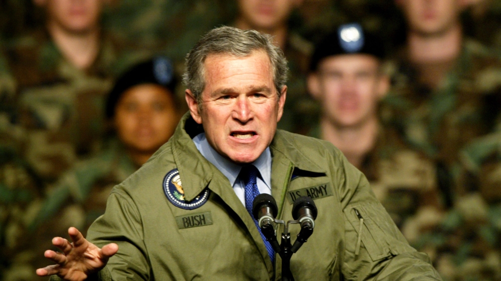 Bush in Army jacket that says "Bush" on one side and "US Army" on the other, at the microphone in the middle of a speech. His face is frozen in a fierce expression. Behind him, blurred, is a group of US soldiers.