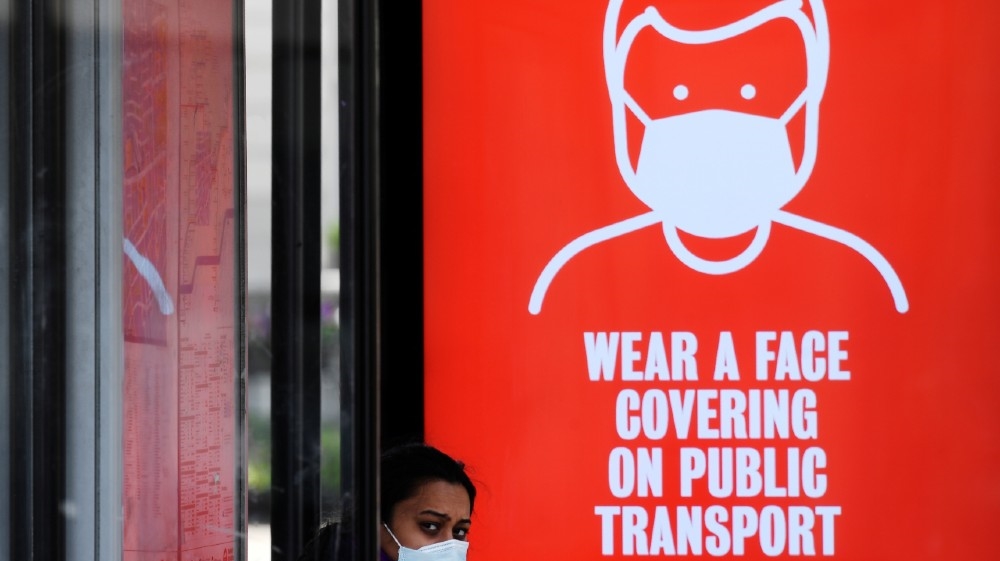 A woman wearing a protective face mask is seen at a bus stop with a public health campaign advertisement asking for passengers to wear masks on public transport