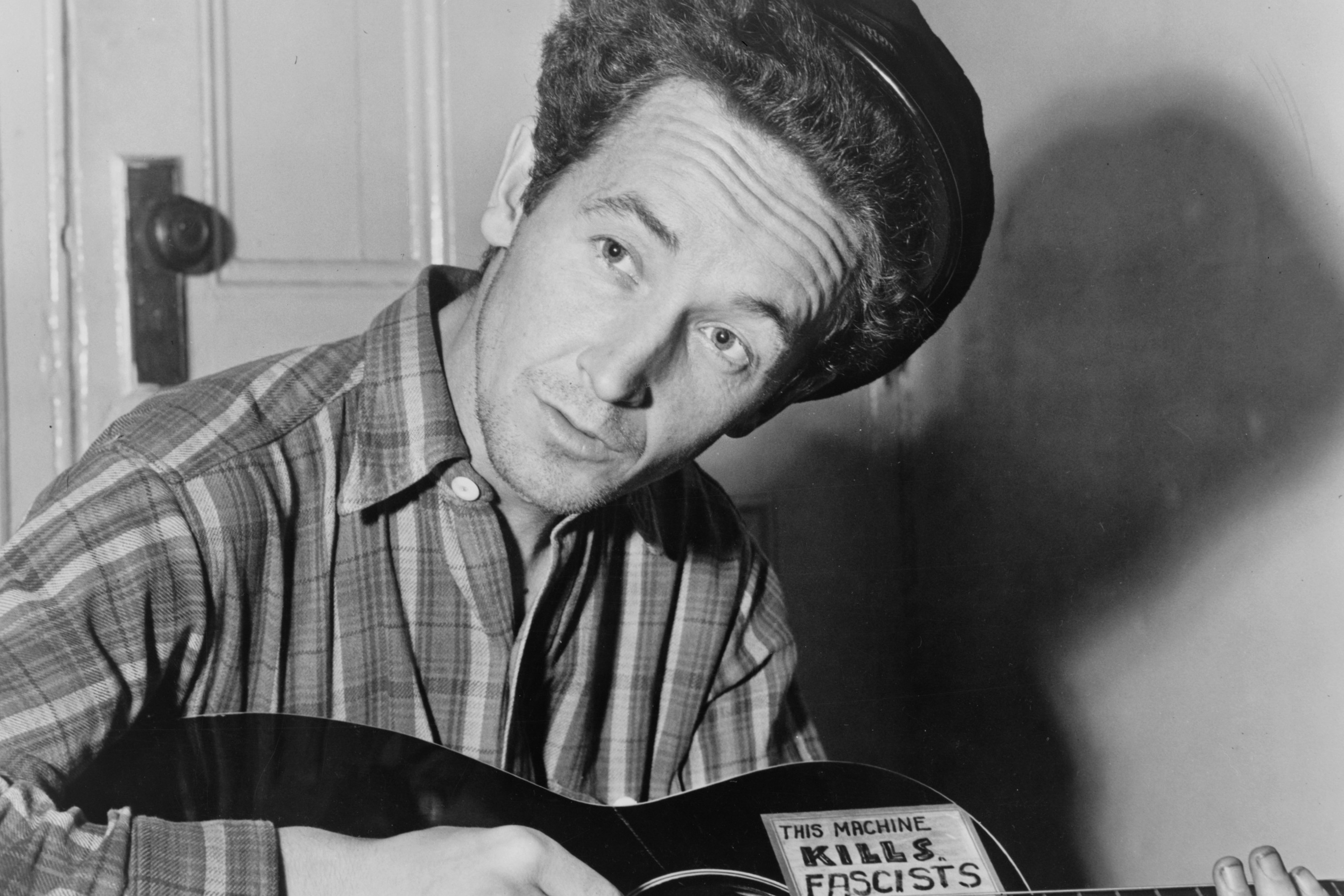 Woody Guthrie is seated playing a guitar that has a sticker attached reading: This Machine Kills Fascists [Library of Congress]