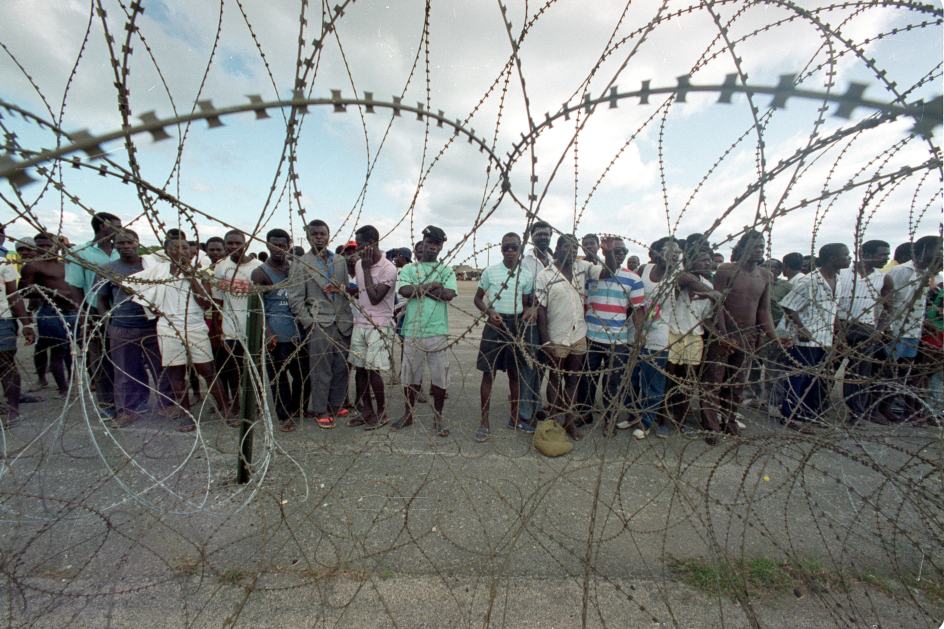 Rows of Haitian refugee men stand behind a razor wire enclosed camp at the Guantanamo Bay Naval Base in Cuba.