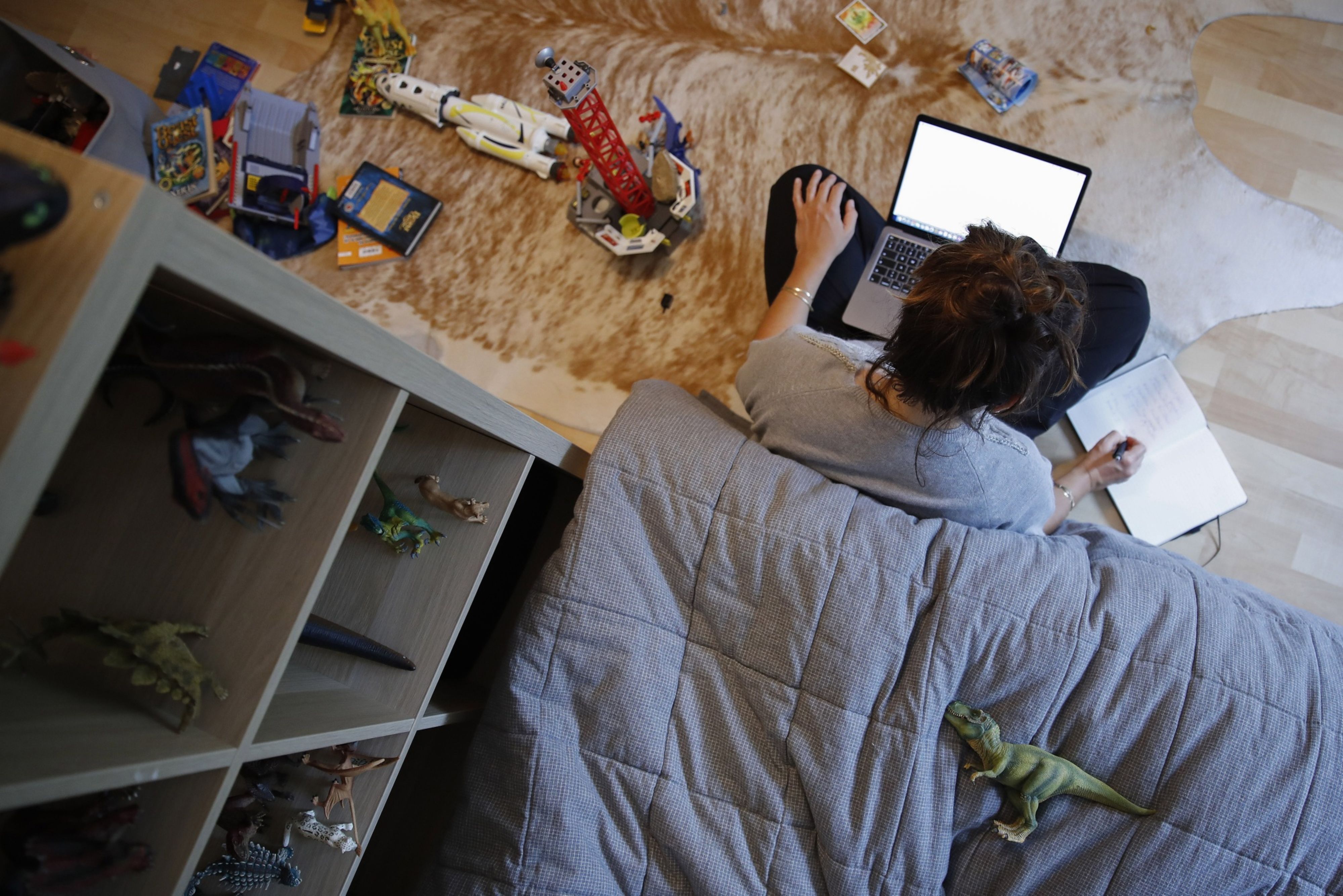 A woman works at a laptop computer on the floor of a bedroom surrounded by children's toys in this arranged photograp