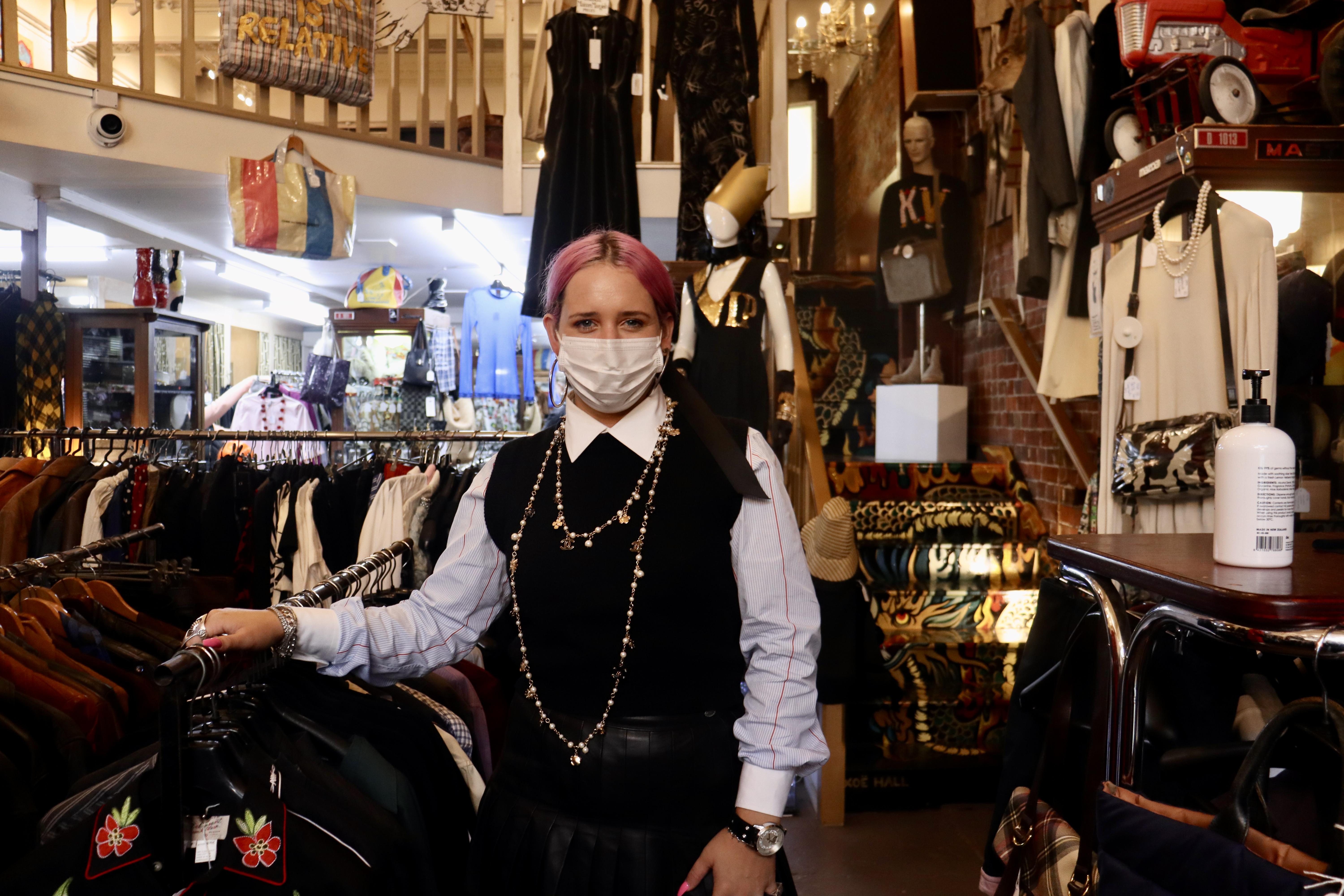 Charlotte Hall in blue shirt and black woollen vest surrounded by vintage clothing in the shop where she works