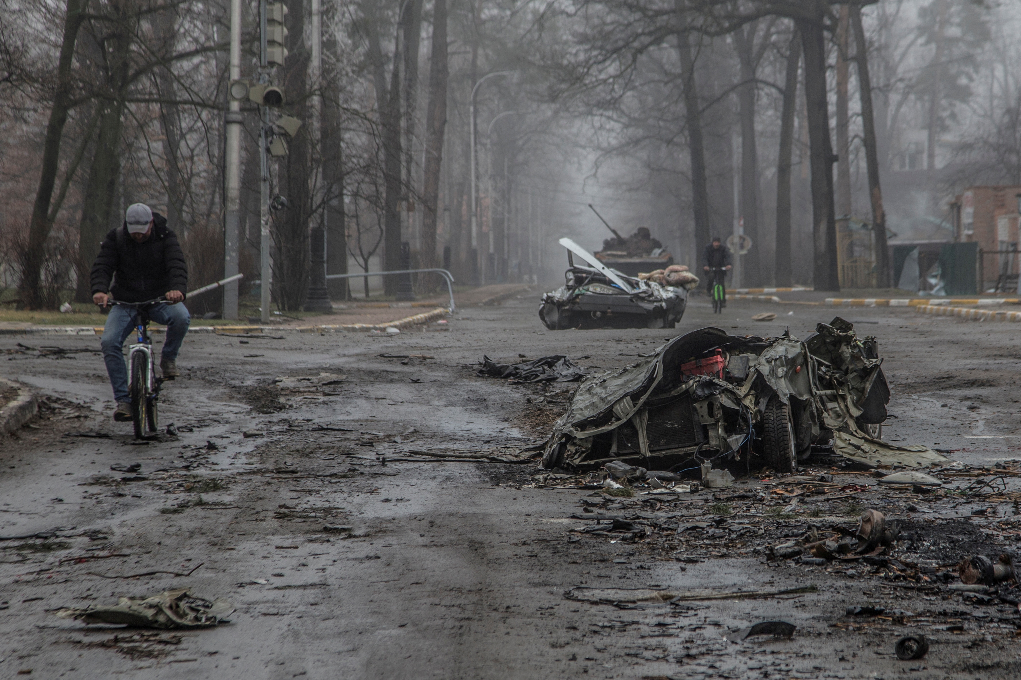 Local residents ride bicycles past flattened civilian cars