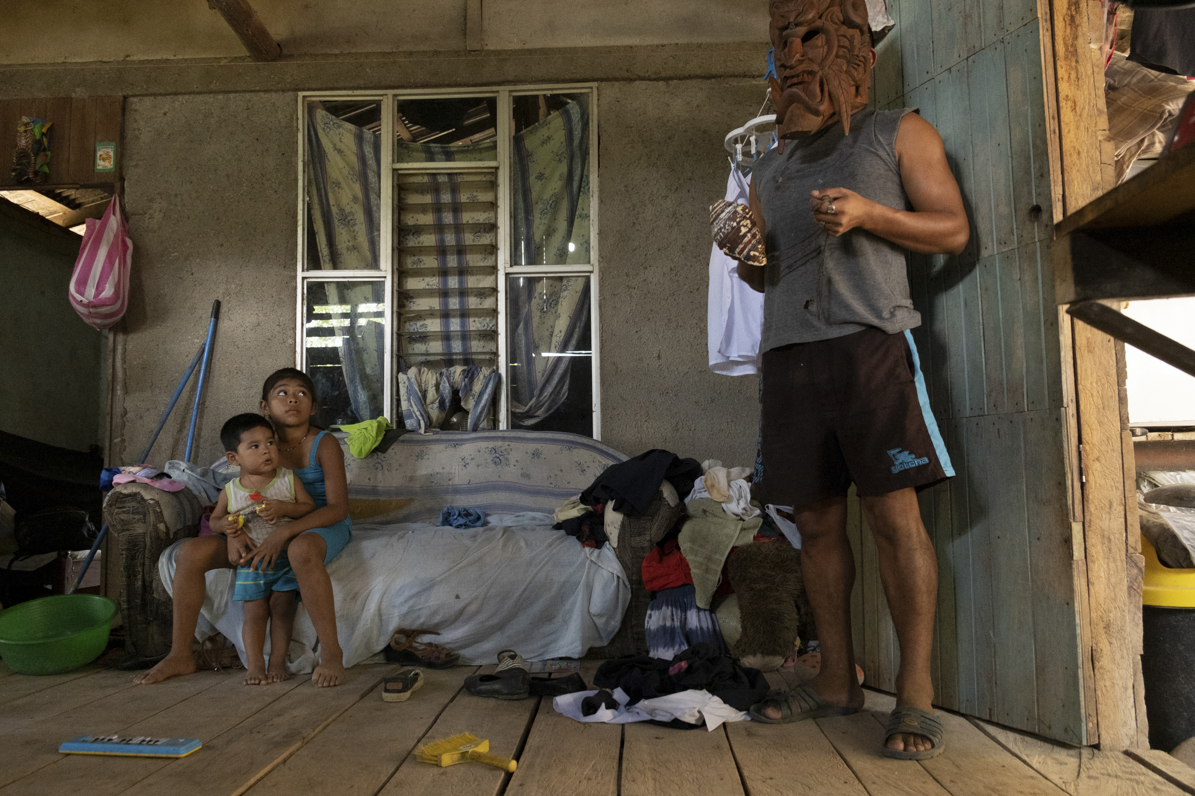 A photo of the inside of a house with two children sitting on a sofa looking at a person wearing a mask standing next to them.