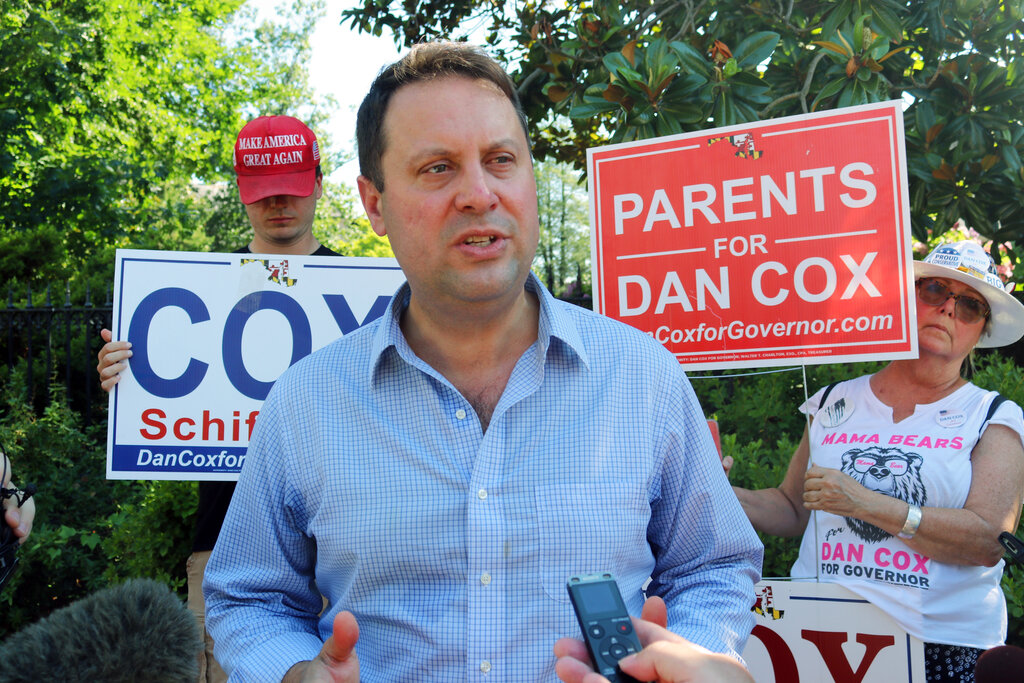 Dan Cox talks to reporters with some supporters holding his campaign signs in the background.