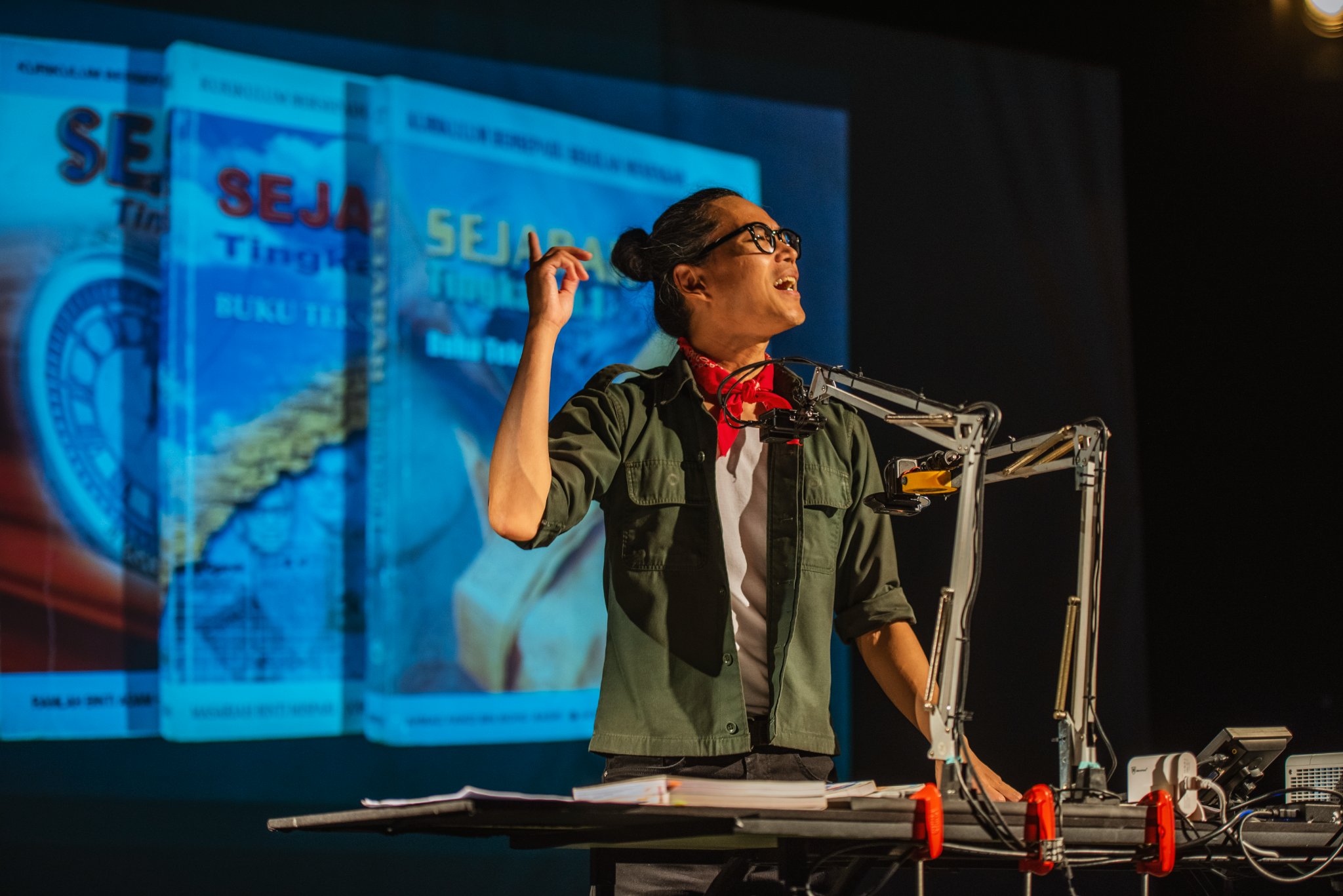 A performer on stage with a video of Malaysian history books behind