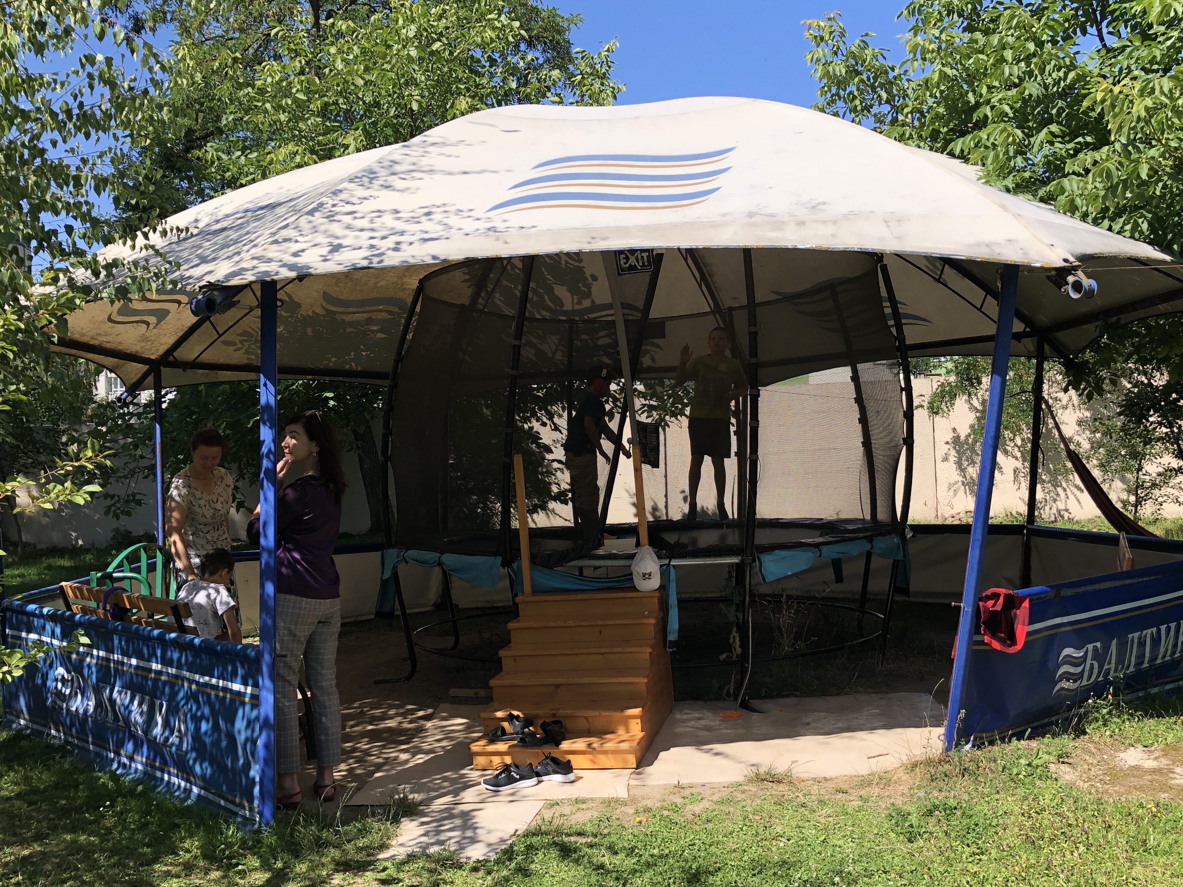 A photo of a Trampoline under a large shade and two people standing and a child sitting on a bench on the side of the shade.