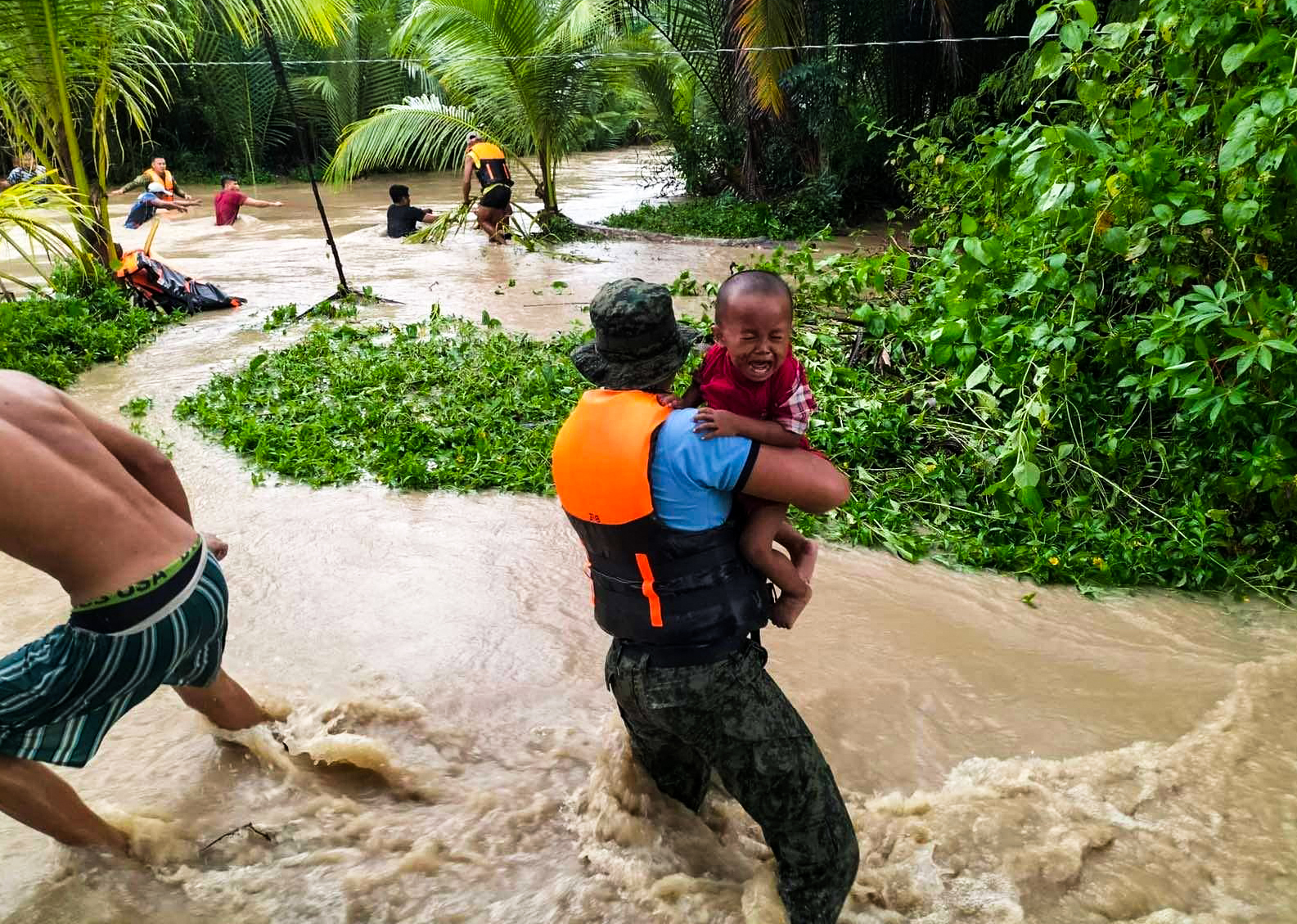 Man holds crying child as he stands in what looks to be fast-running flood waters. In the background, people try to cross the water holding onto a rope.