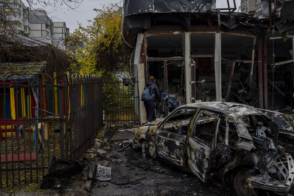 Two people peering inside the shell of a building. A destroyed car is behind them. There is debris all over the ground.