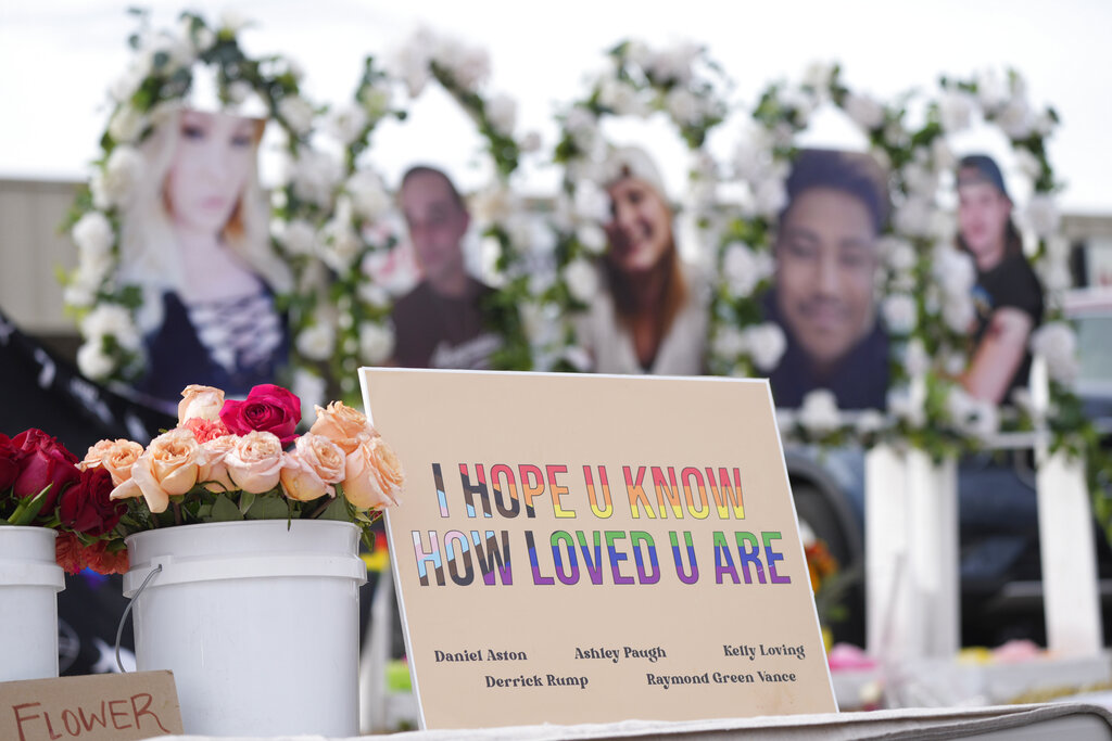 A memorial sign saying 'I hope u know how loved u are' next to a bucket of roses, with the faces of victims in the blurred background