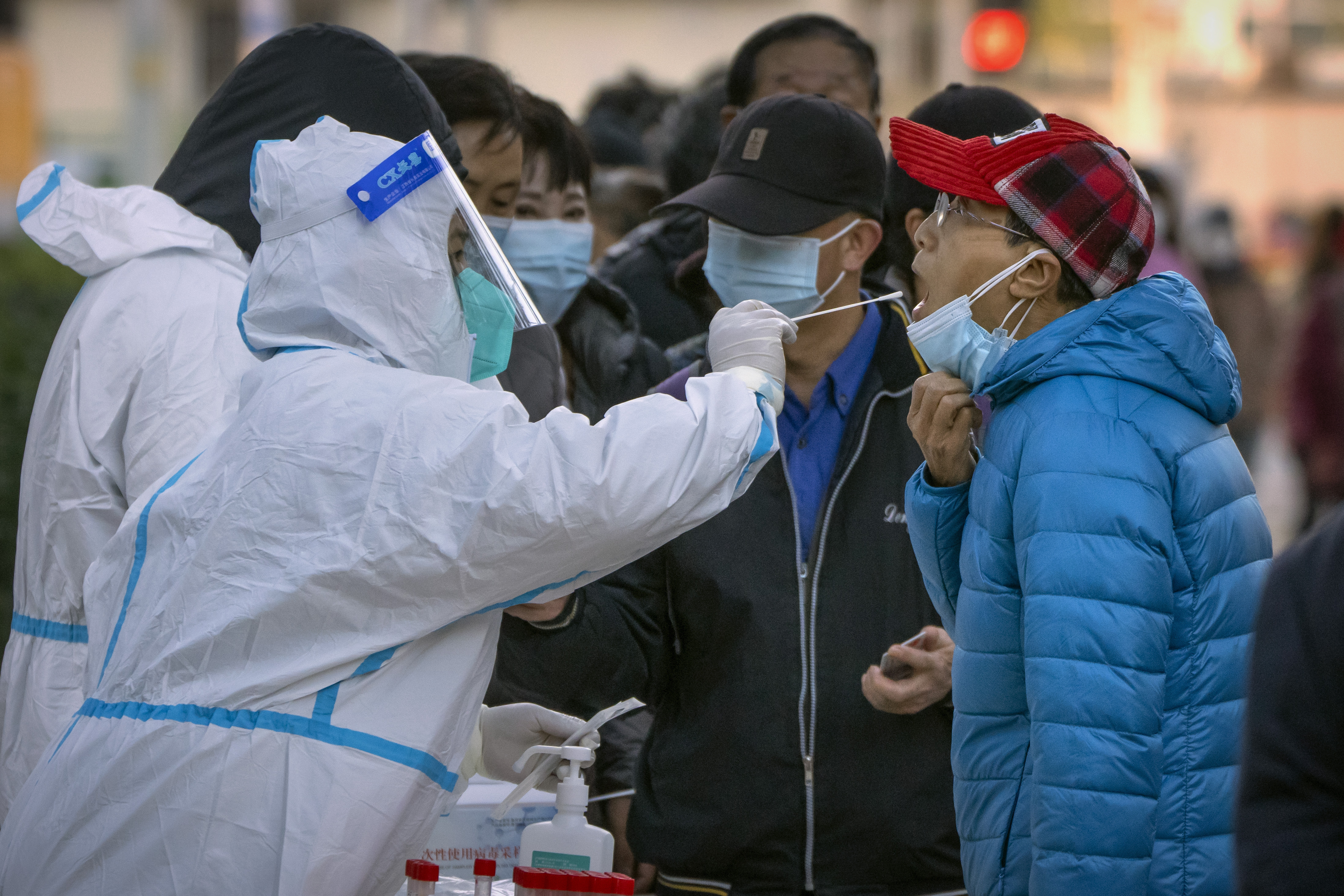 Woman in full protective gear holds a swap next to a man's open mouth. Others wait behind with masks on..