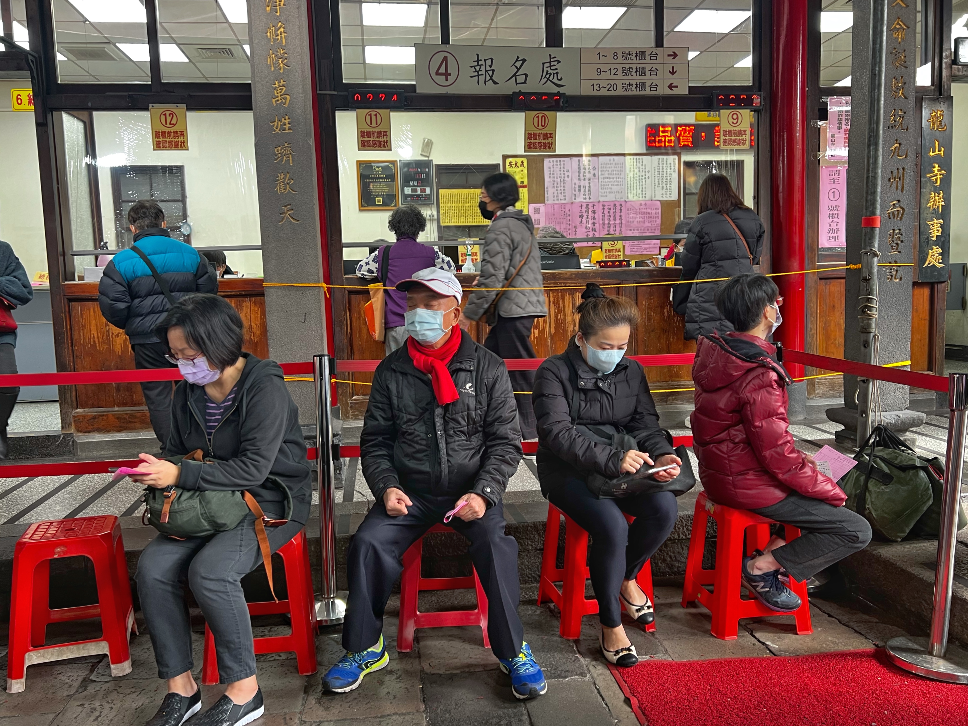 People waiting to an tan sui or pray to other gods at the temple. They are sitting on red plastic stools. Behind them are counters where they pay for lamps and candles