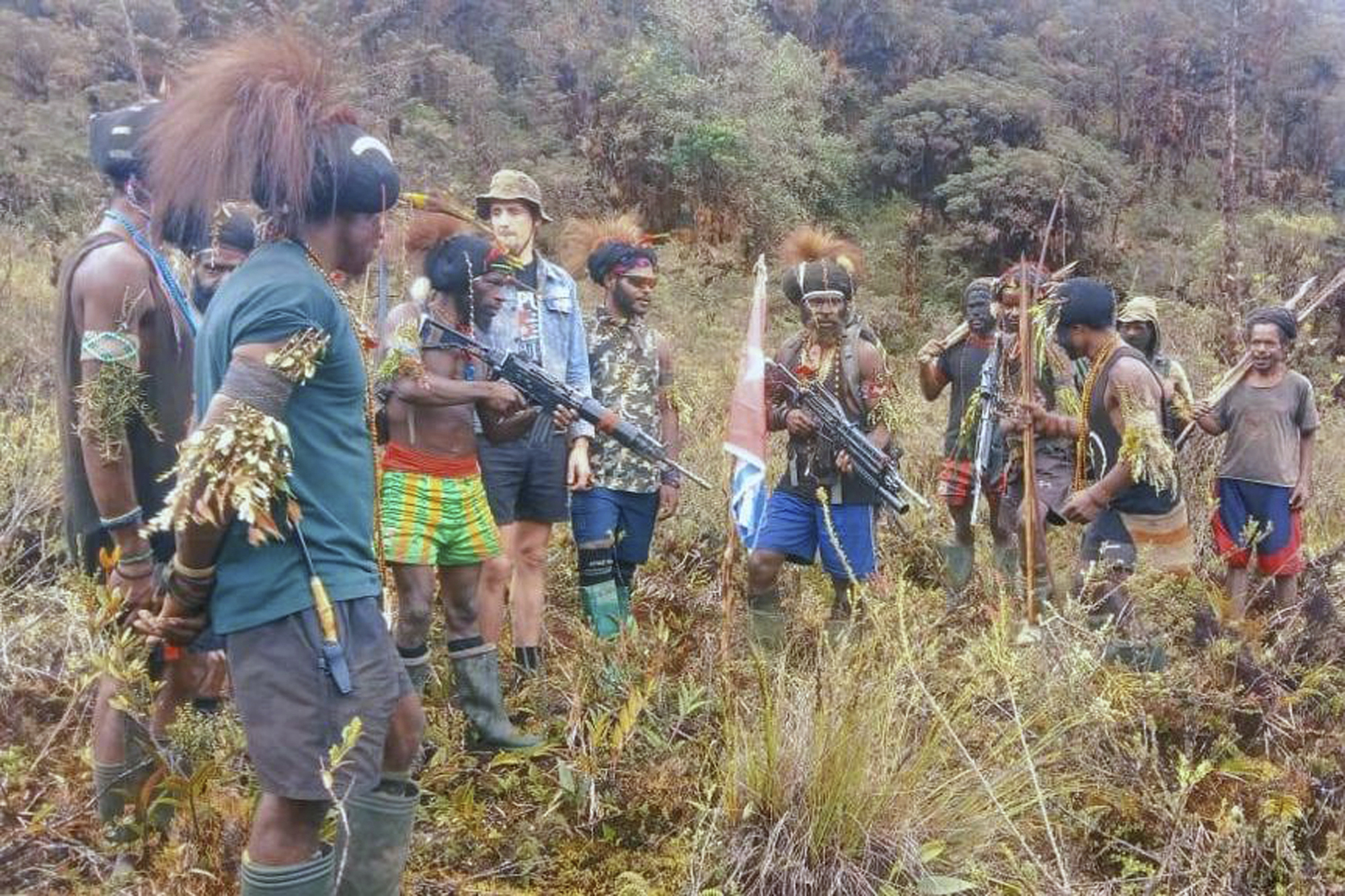 A man said to be Phillip Mehrtens in a line with armed West Papua rebels, The rebels are in shorts, t-shirts and wellington boots. Some are wearing traditional hats with plumes and have leaves and grasses tied to their biceps. There is a West Papua independence flag behind them.