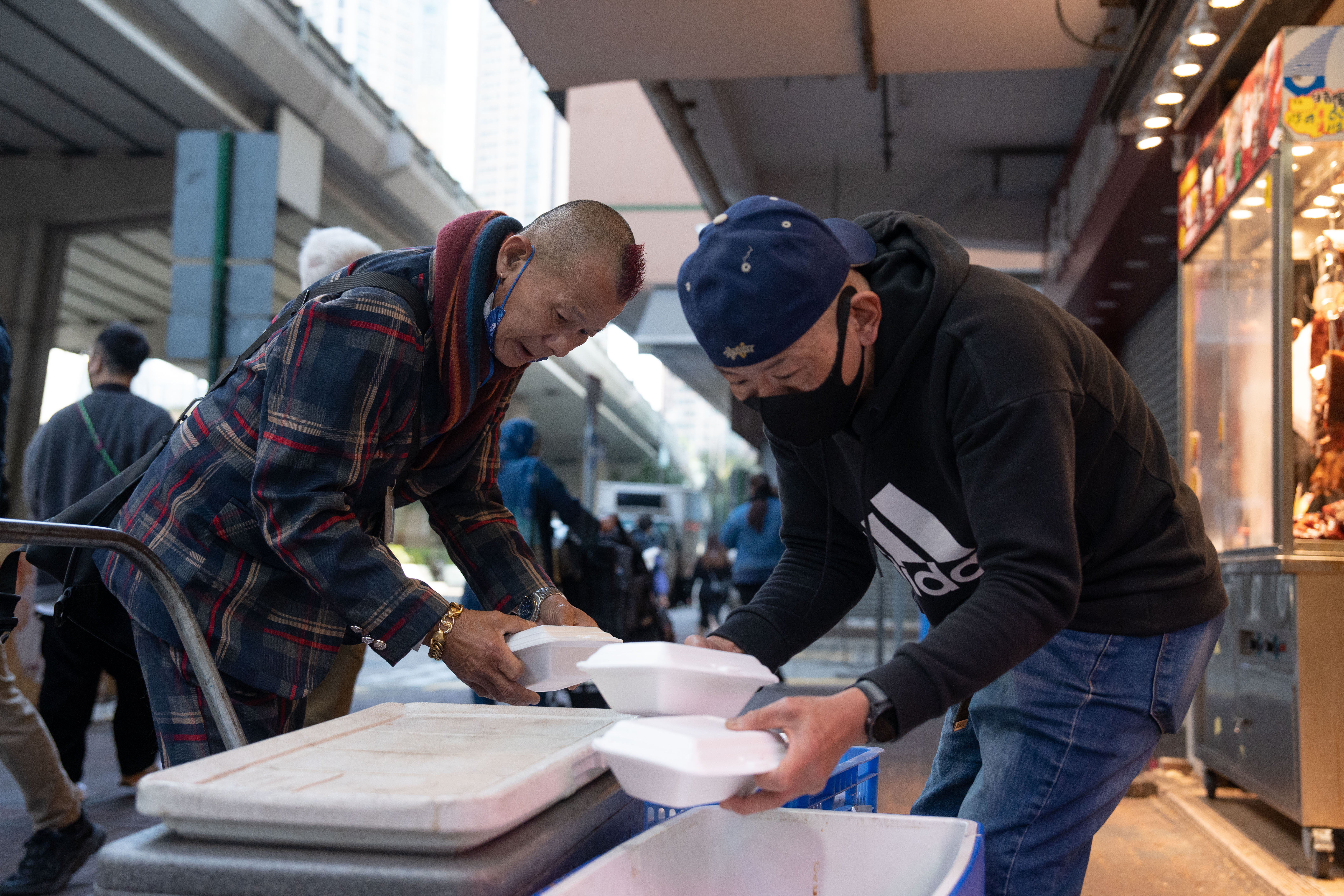 Ah Lik organises food parcels for homeless people with another HKImpact worker. They are piling the packets onto a trolley, outside a restaurant.