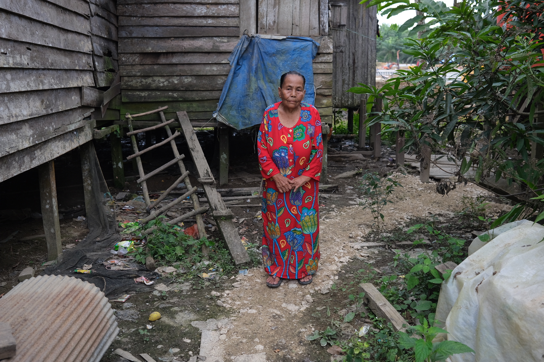 Sernai in a long red dress with a striking blue and green formal pattern. She is standing outside her house and construction can be seen going on in the background through the foliage.