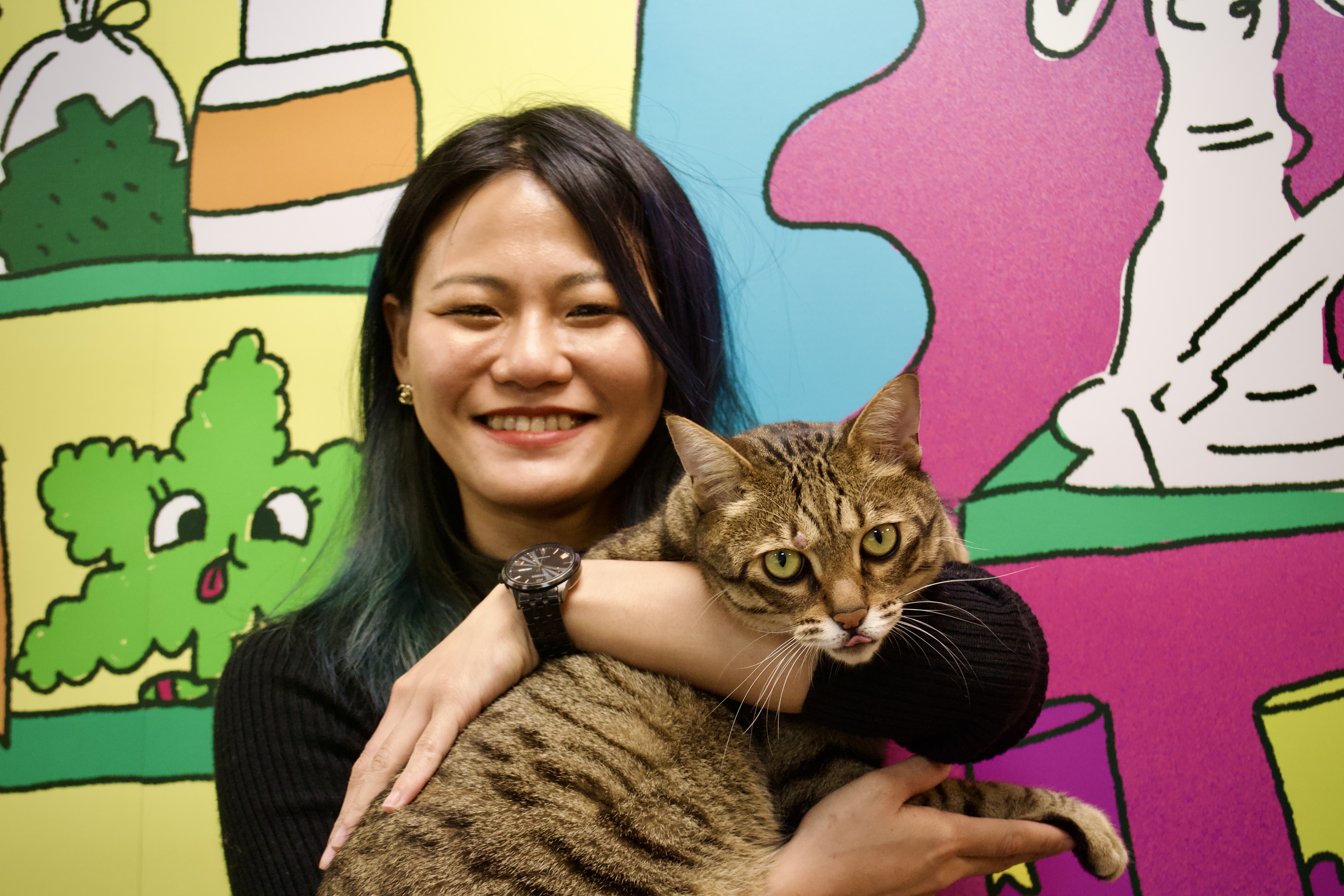Zoe is smiling, wearing a black, long-sleeve top and black watch and holding a cat. She is standing against a colourful mural.