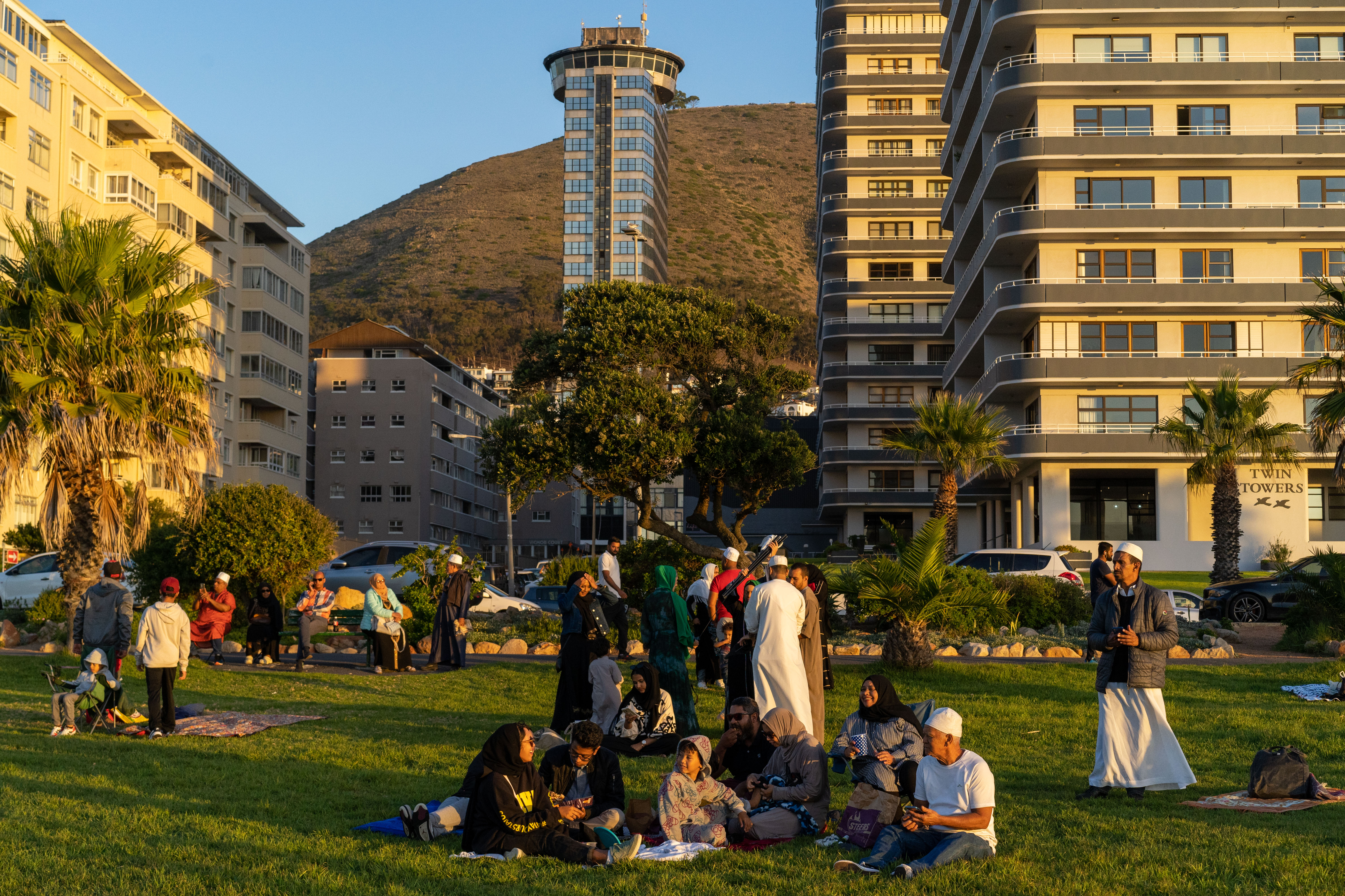 A photo of a group of people sitting in a grassy area in a park with several other people walking around.