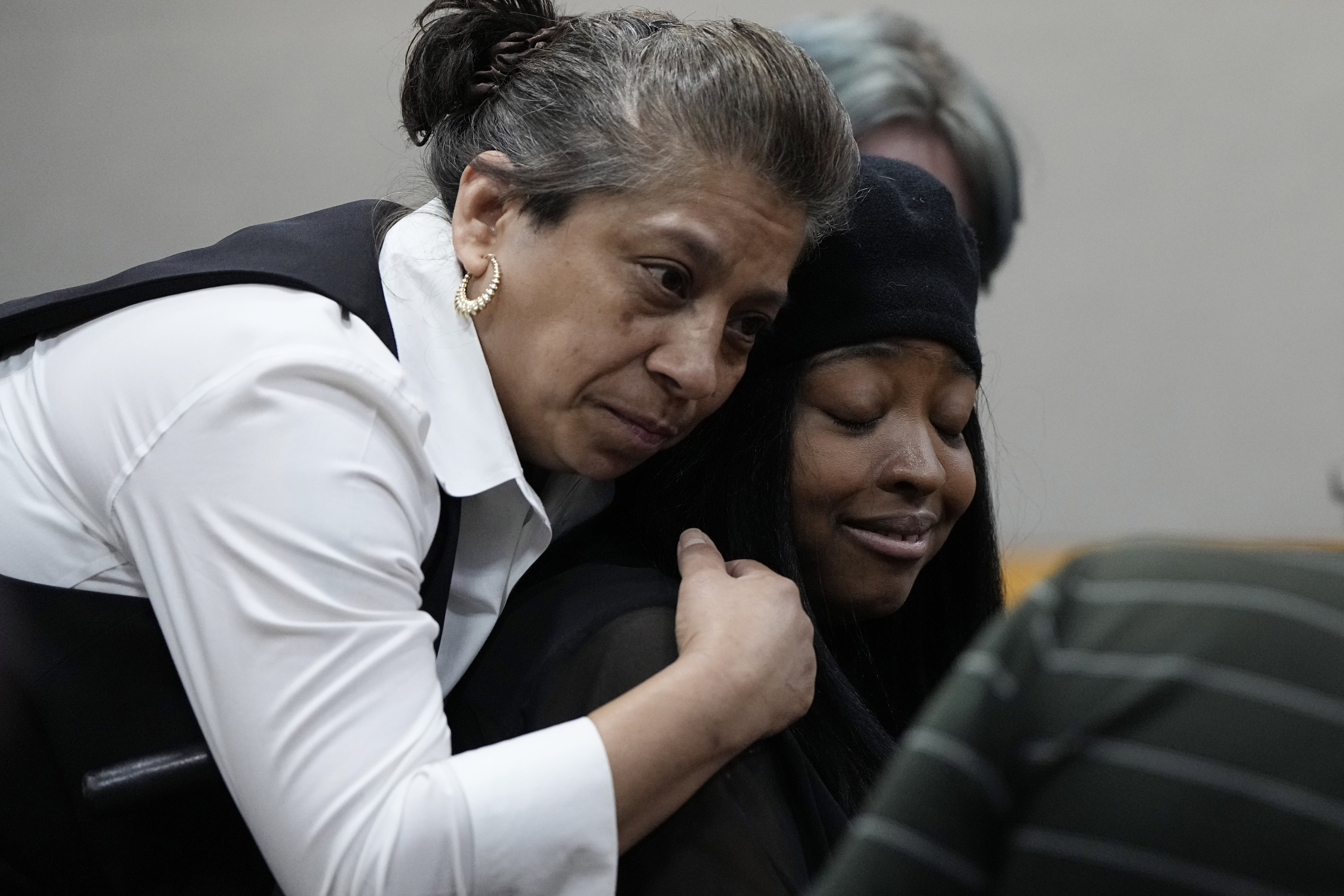 A woman leans over to hug a second woman from behind as she cries in the courtroom