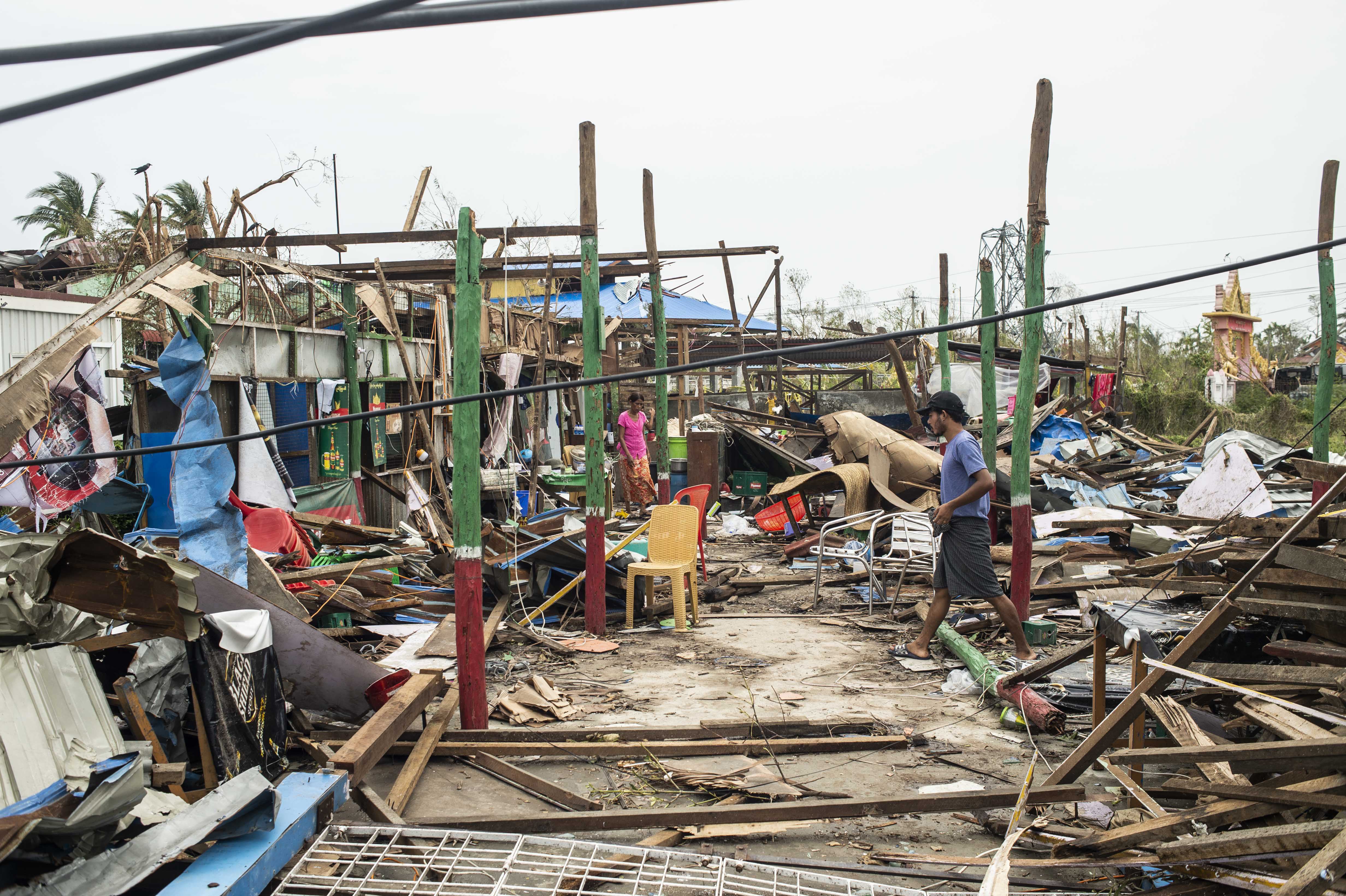 Buildings left without walls or roofs in Sittwe. There are piles of debris between posts that once held up buildings. Two people are walking around the ruins.