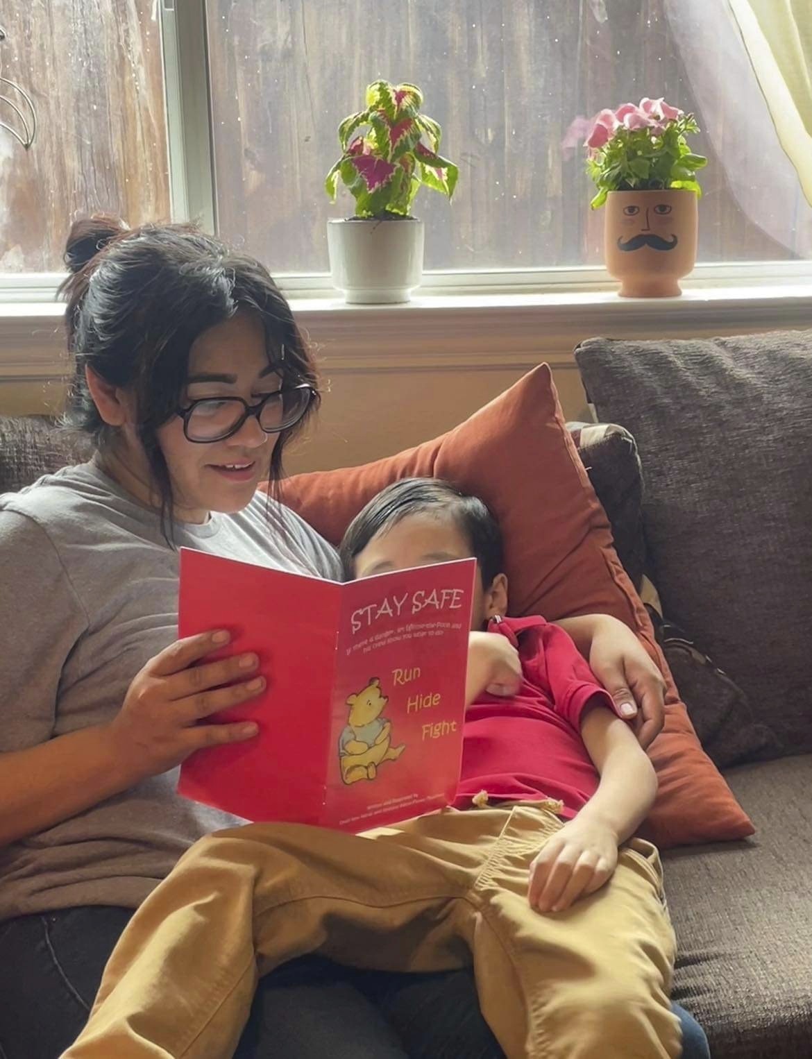 Cindy Campos reads the book "Stay Safe" to her son in Dallas. She is on the couch, wearing glasses and holding the book up. The book is red and has a yellow Winnie the Pooh on the cover with the word STAY SAFE in playful text at the top. Cindy has her arm around her son who is lying back against a pale red cushion and has his legs across his mum's lap.