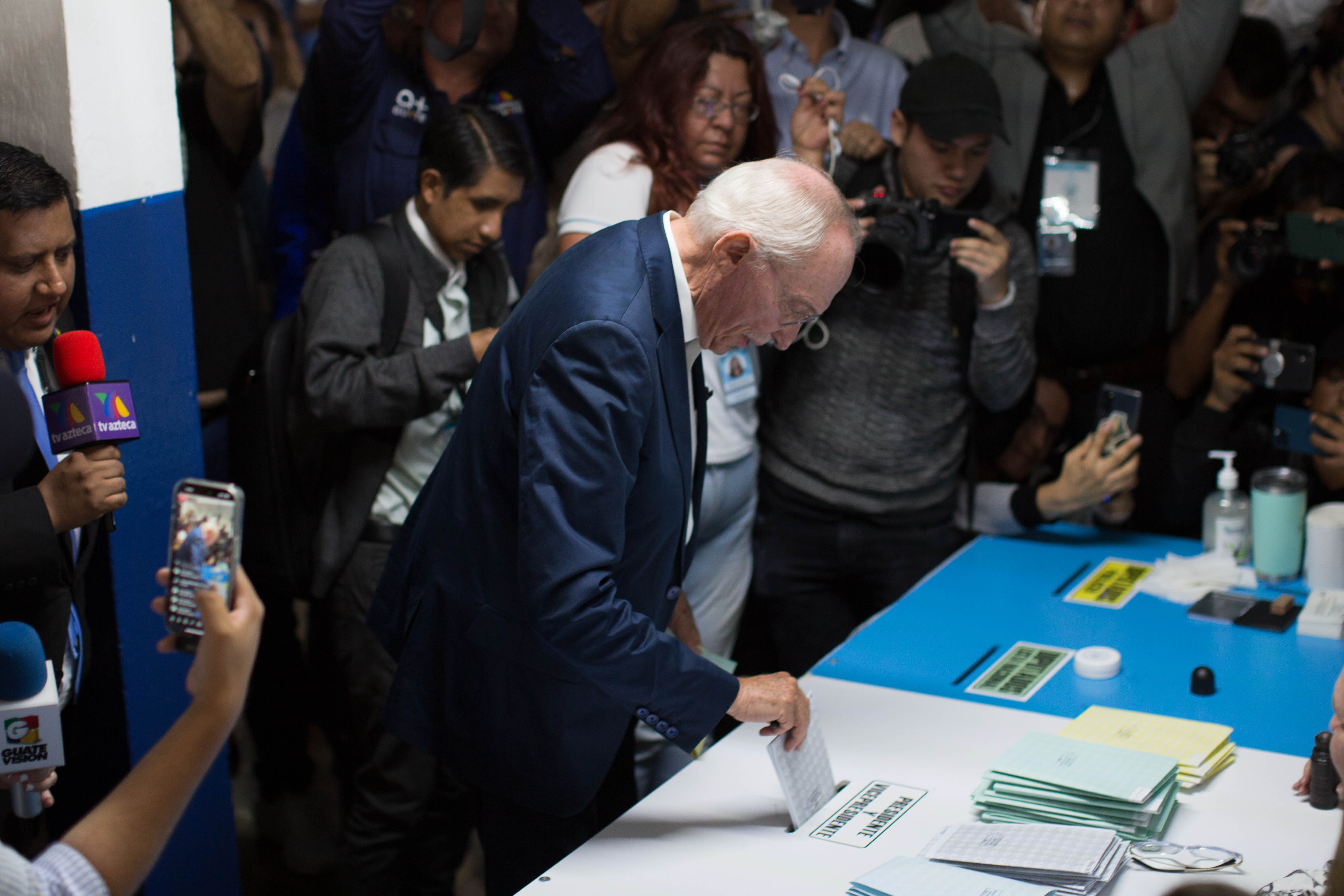 A man in a suit leans over a large white ballot box, placing a sleeve of paper in an opening along its top.