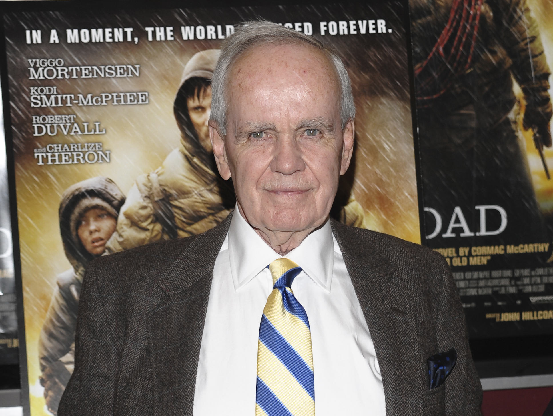 A man in a suit and striped tie stands in front of a movie poster for The Road.