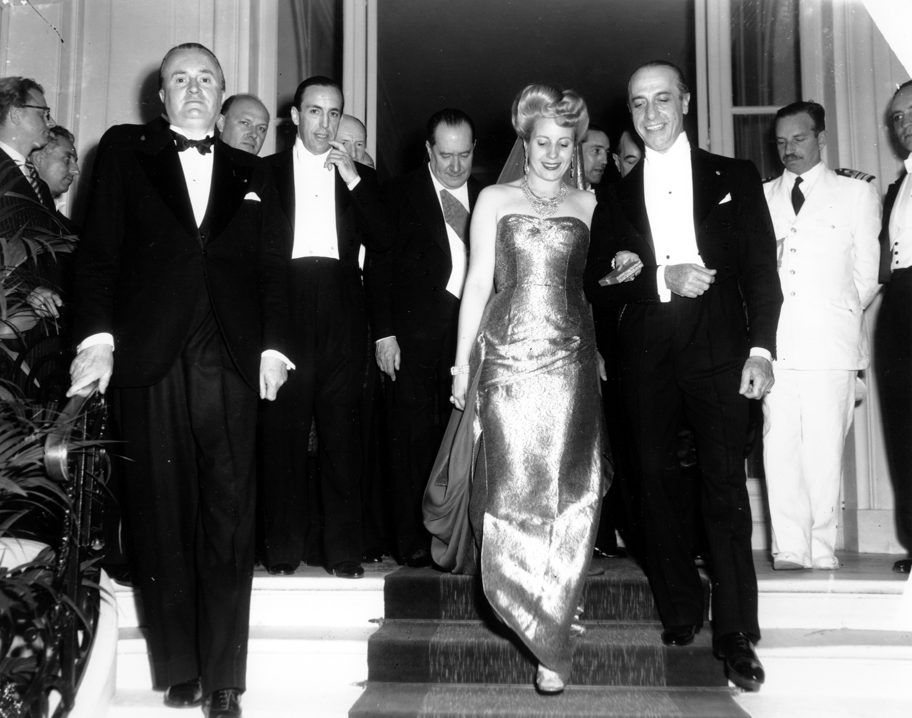 A woman in a shiny gown walks arm-in-arm with a man in a white tie as they travel down a set of steps
