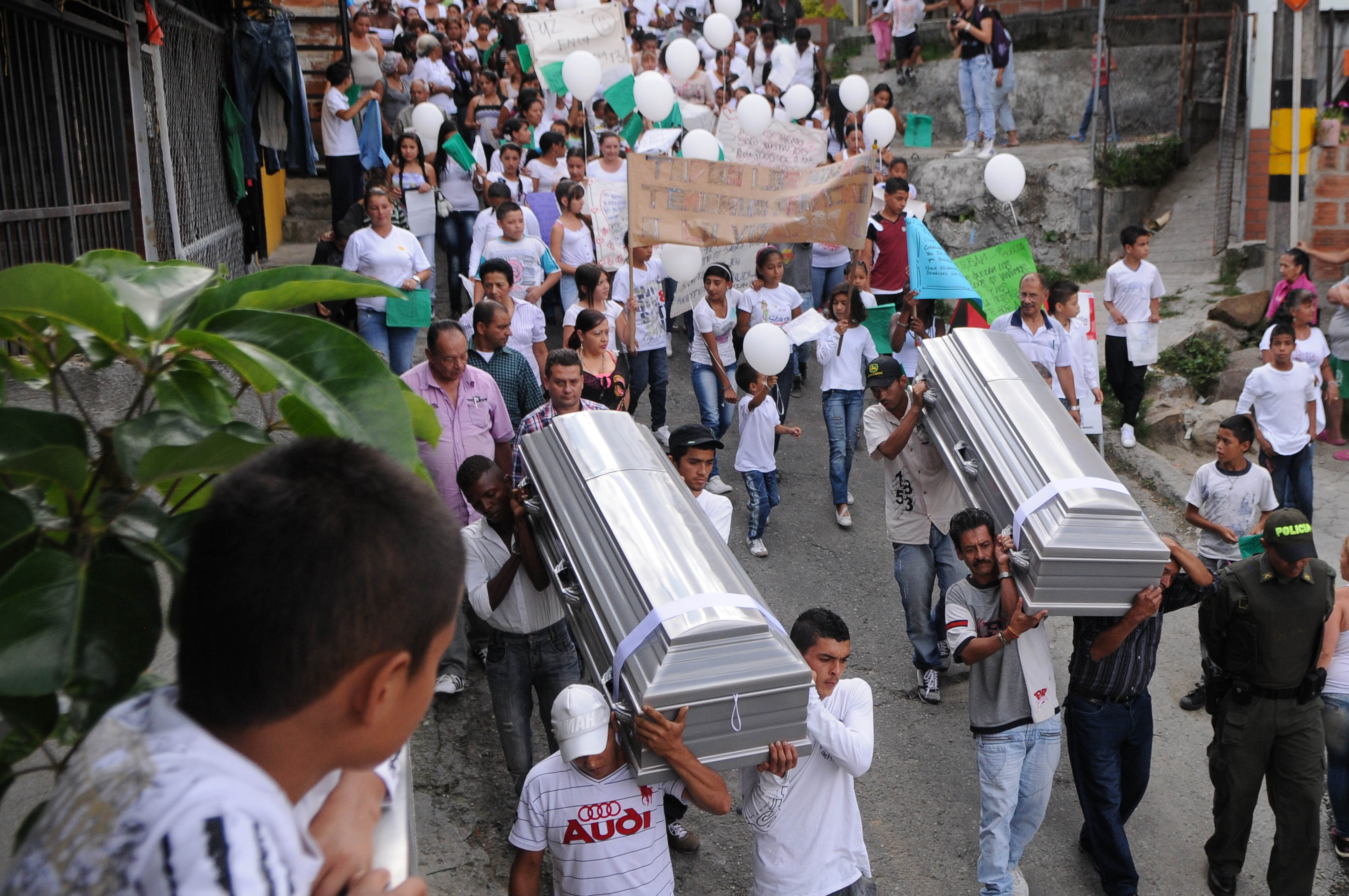 A boy peers over a balcony and onto a street, where a group of mourners carry two small silver coffins.