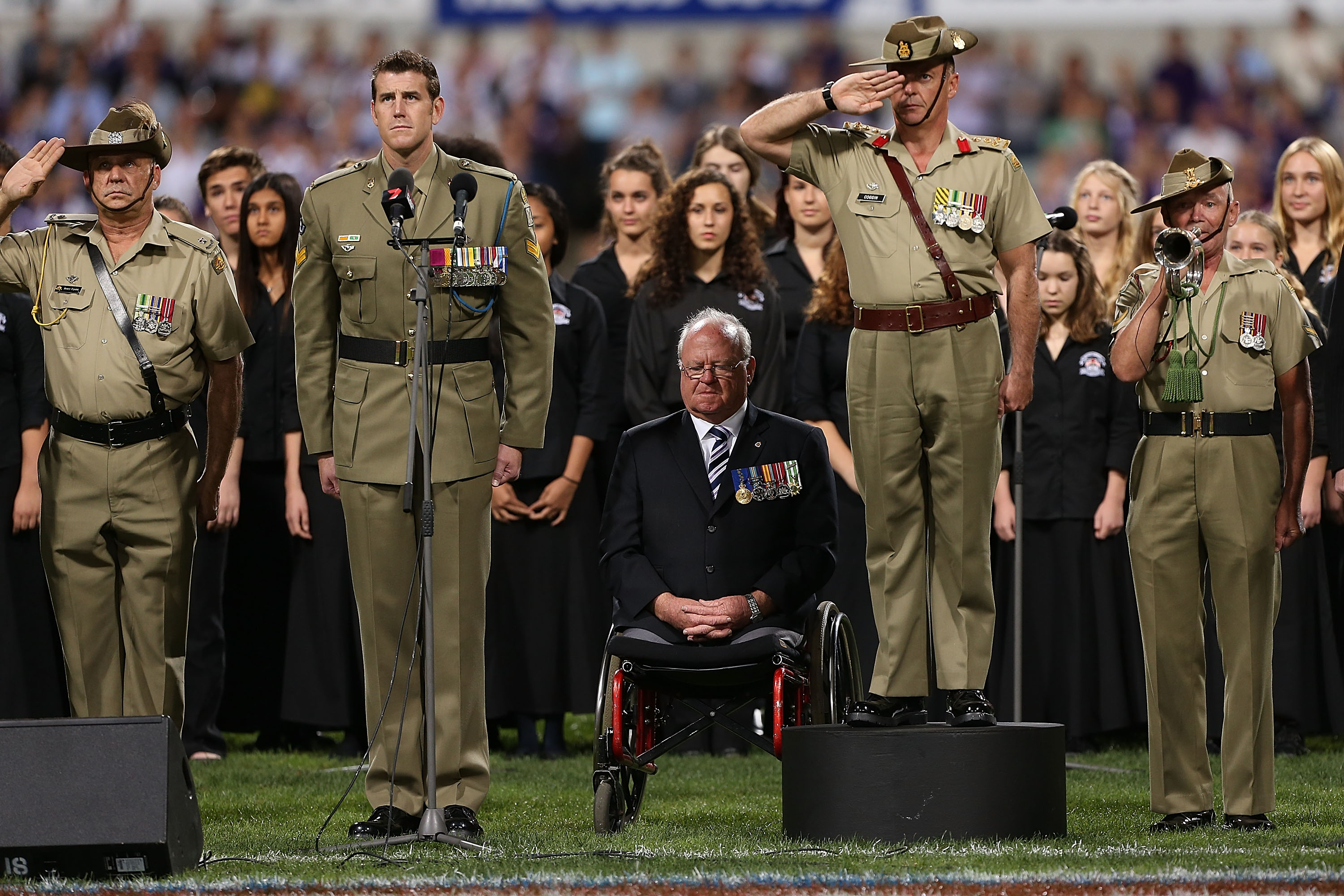 Ben Roberts-Smith standing to attention ahead of an AFL game in Perth in 2013 as the last post is played. He is in uniform. There is an elderly veteran in a wheelchair next to him, other soldiers around him saluting, and a group of young women behind.