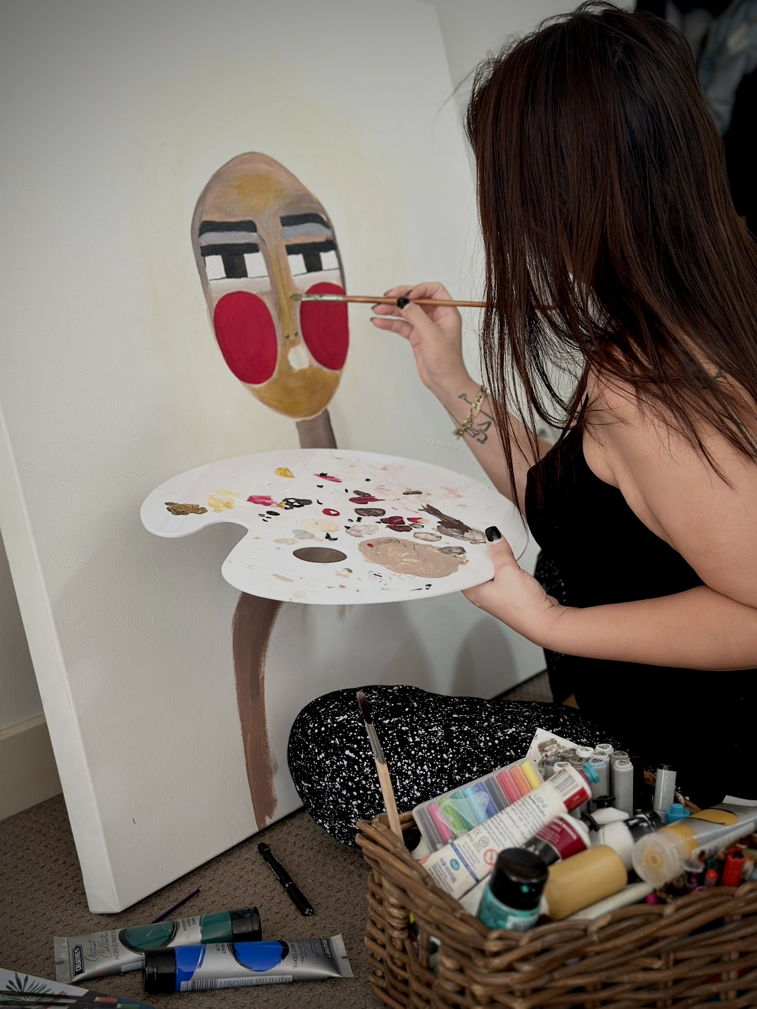 Maya working on one of her paintings. It shows a figure with bright red cheeks. She is holding a palette in her left hand as she works on the canvas.