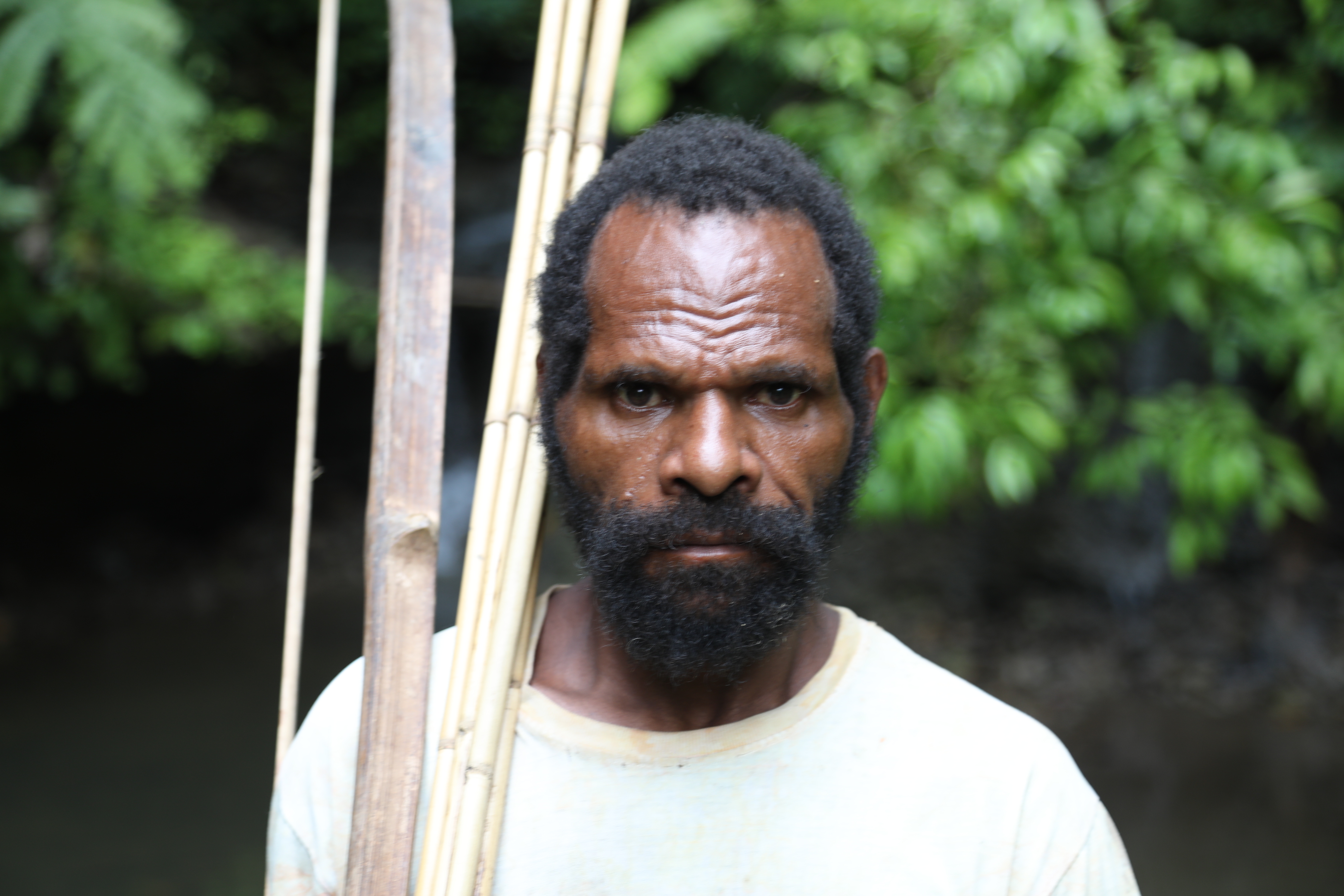 A portrait of Josep Ogoney. He is wearing. white T-shirt and has some bamboo on his shoulder. He is in the foresy