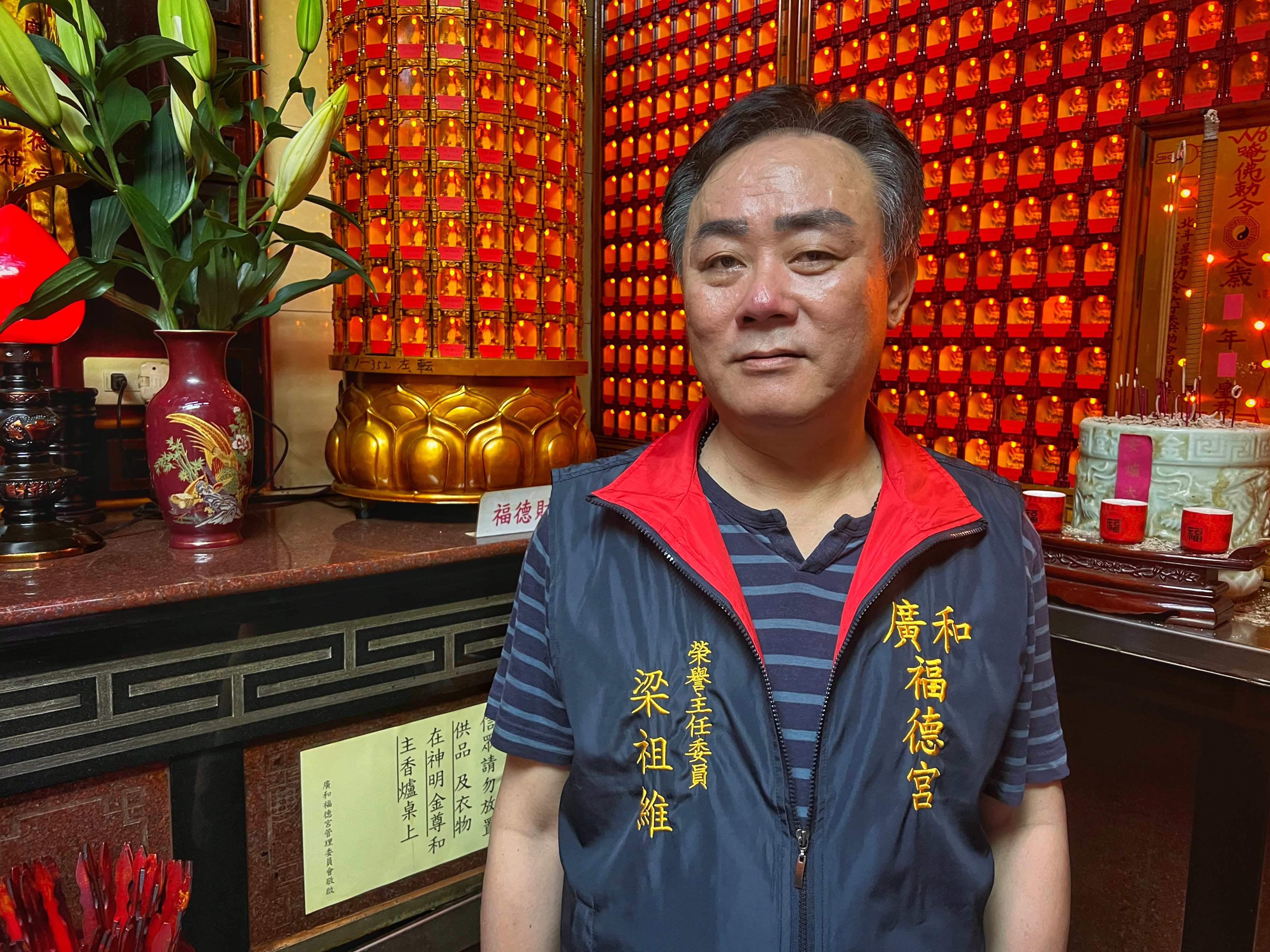 Liang Tsu-wei, honorary chairman of the Guanghe Fude temple. He is standing against a red wall in the temple. He's wearing a blue striped polo shirt, a sleveless jacket in blue with Chinese characters written in yellow.