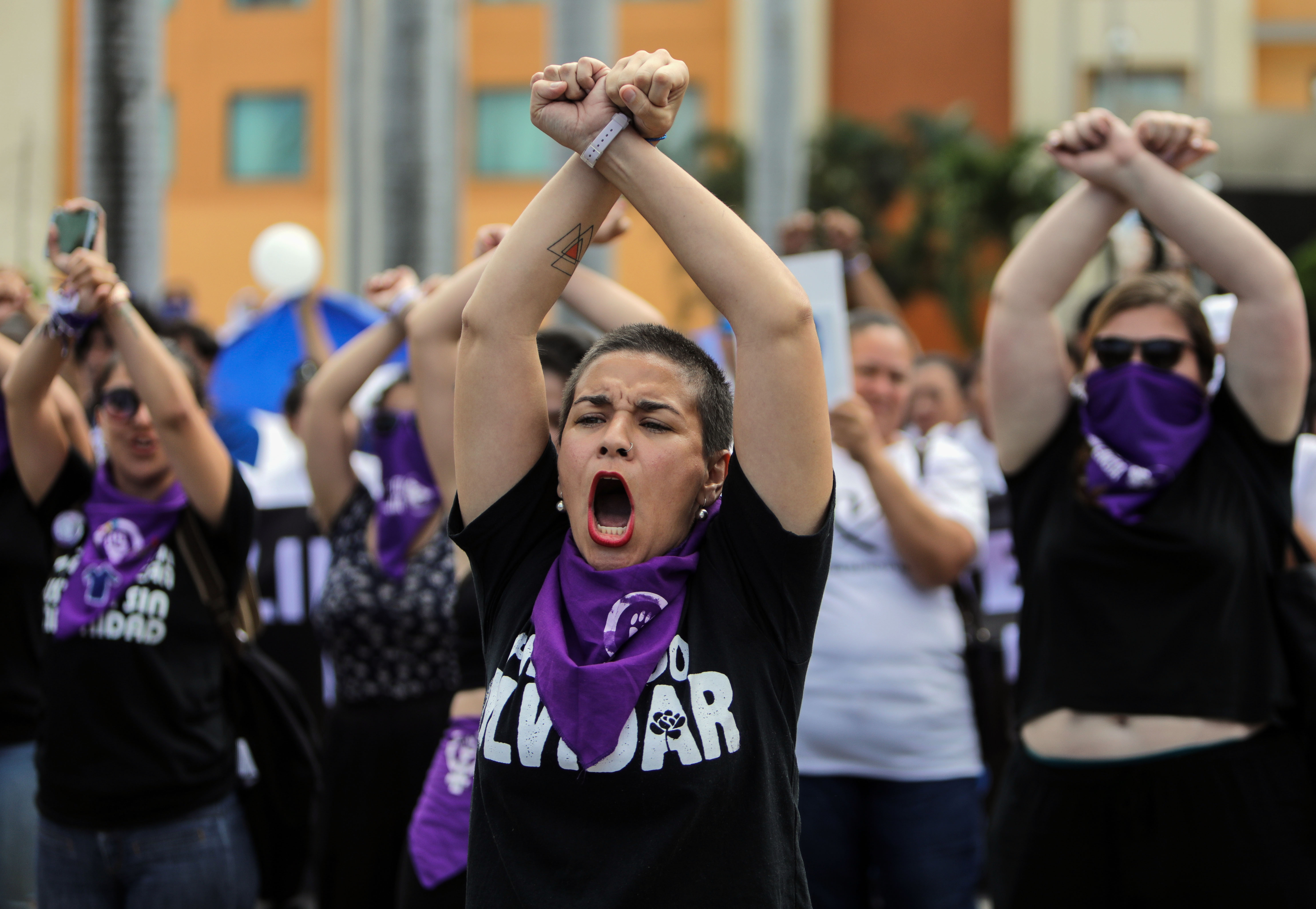 A woman with a purple handkerchief around her neck raises her arms and crosses them above her head, as part of a protest with other activists