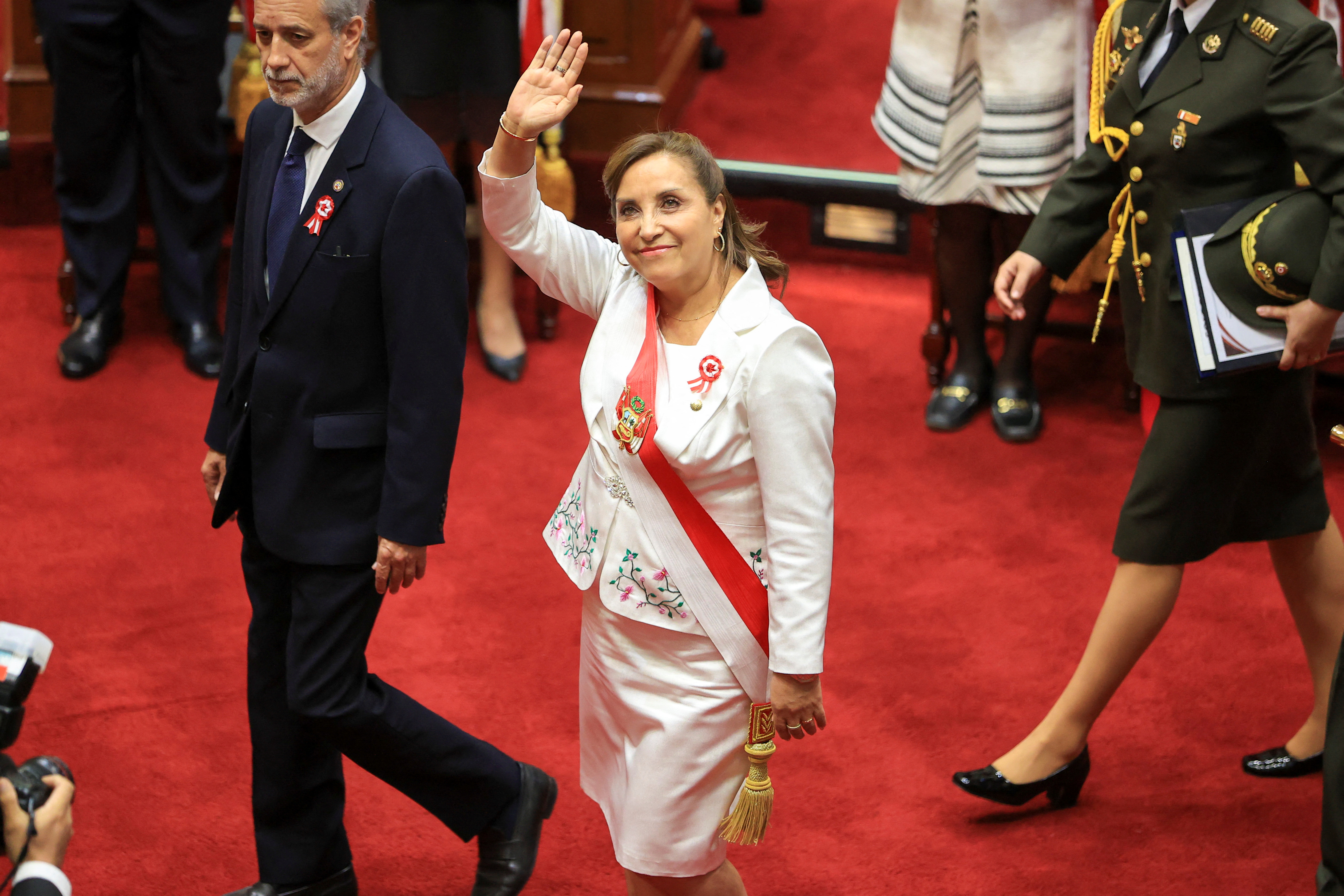 A woman waves up to a balcony as she walks across a red carpet in Peru's congress, wearing a presidential sash.