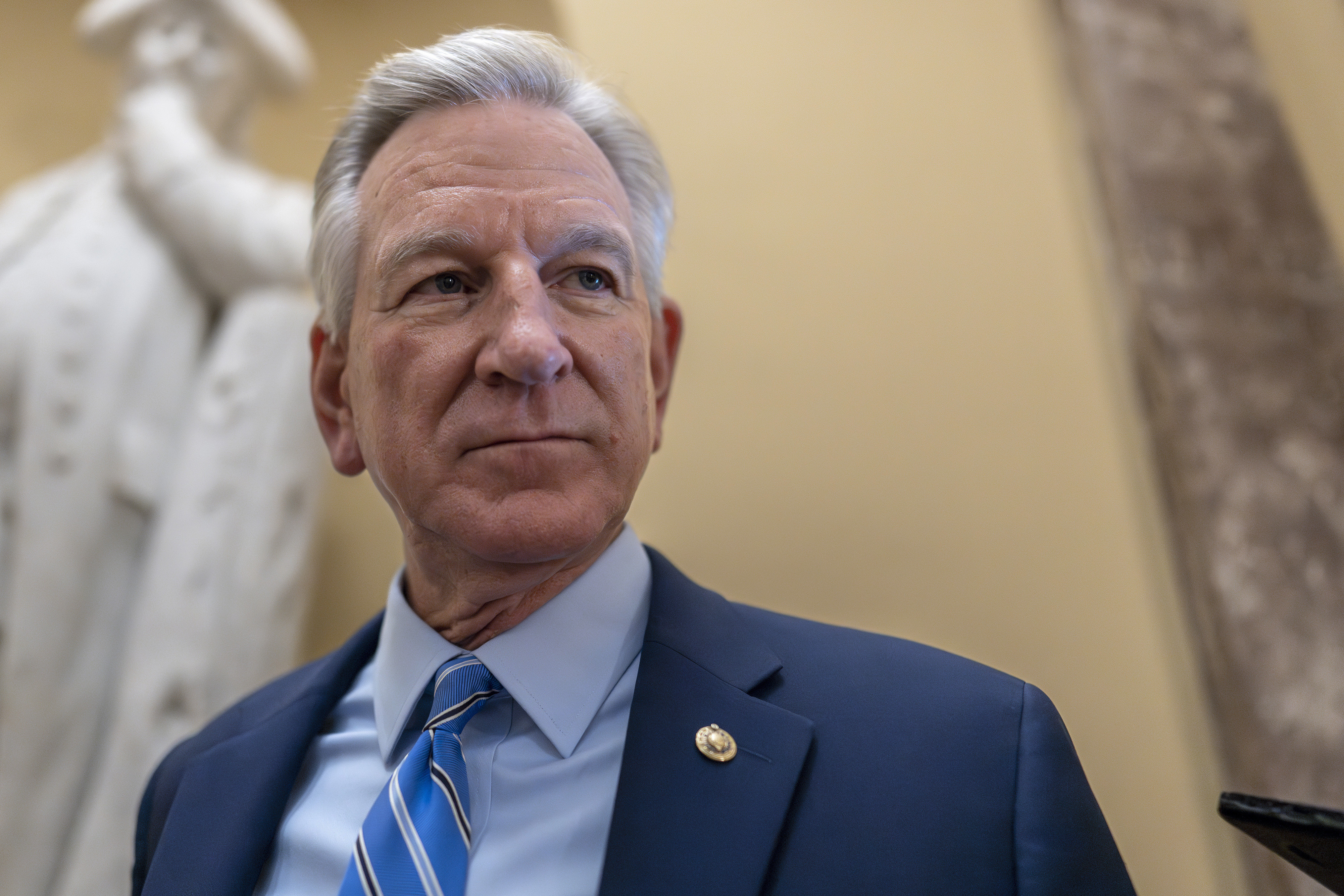 A man, inside the Capitol, stands in front of a marble statue in a blue suit and tie.