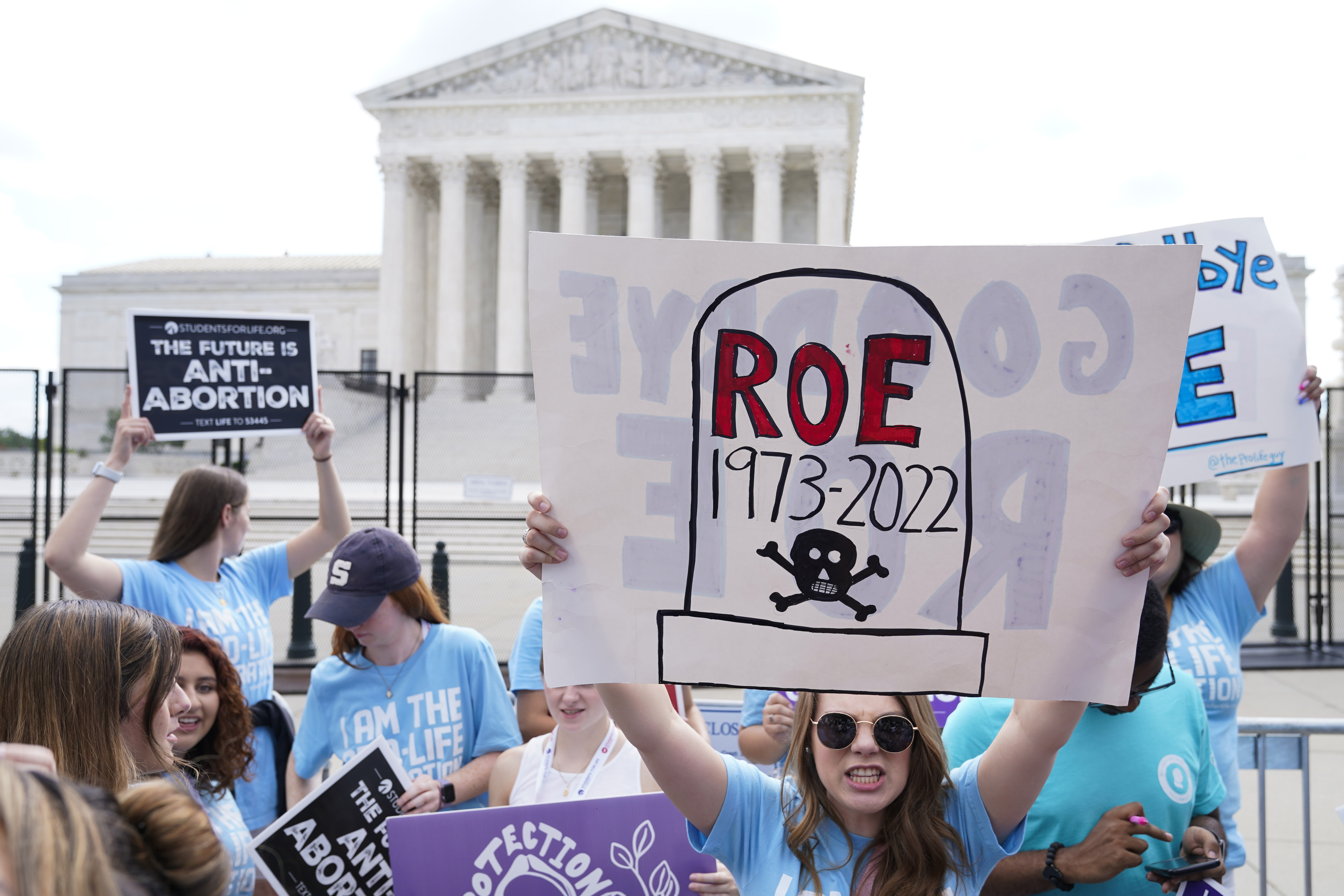 A woman holds up a hand-drawn poster that depicts a tombstone with the words "Roe 1973-2022" written on it. The Supreme Court is behind her.