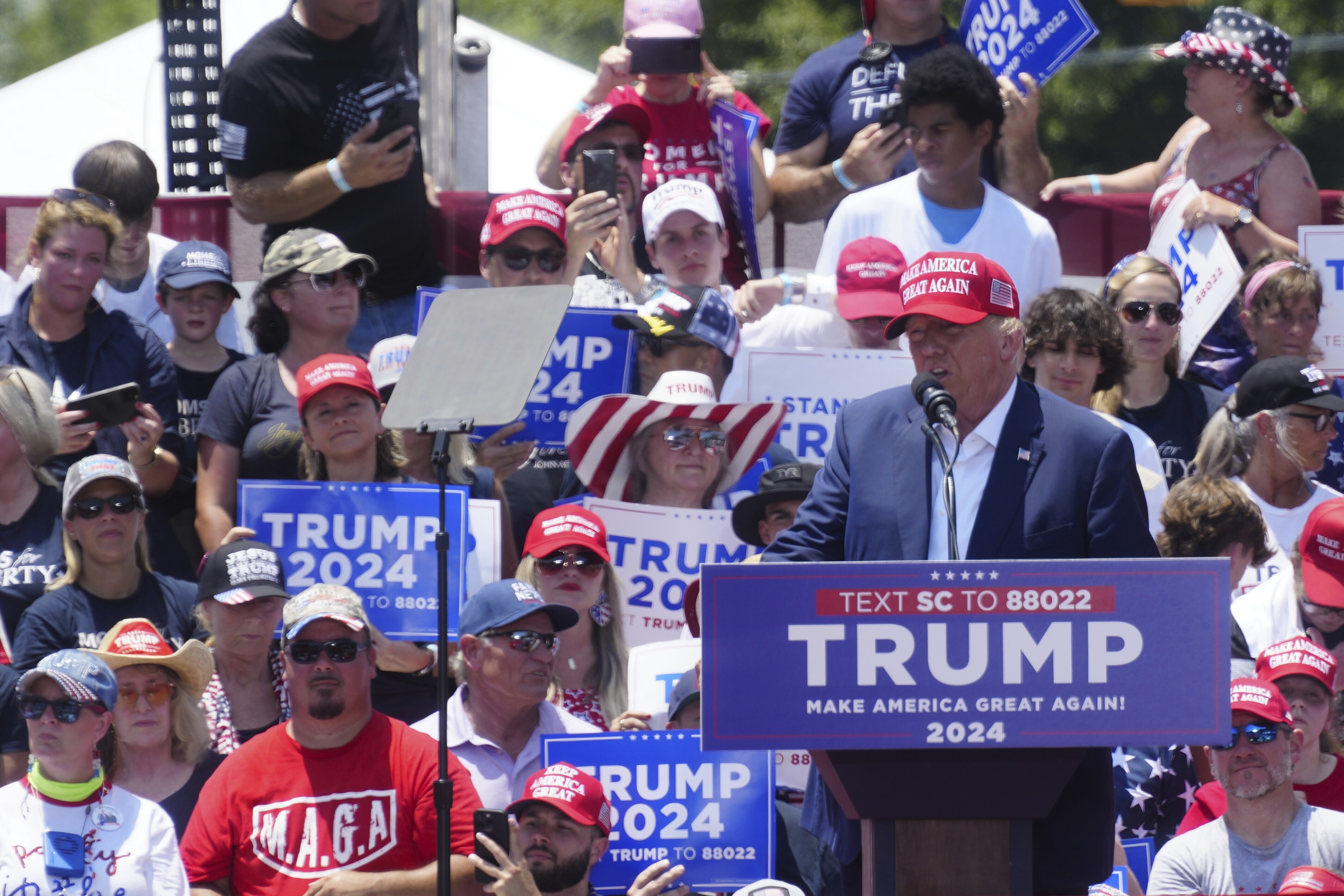 Trump, in a red baseball cap, stands behind a podium with his name on it. A crowd of supporters stands behind him.