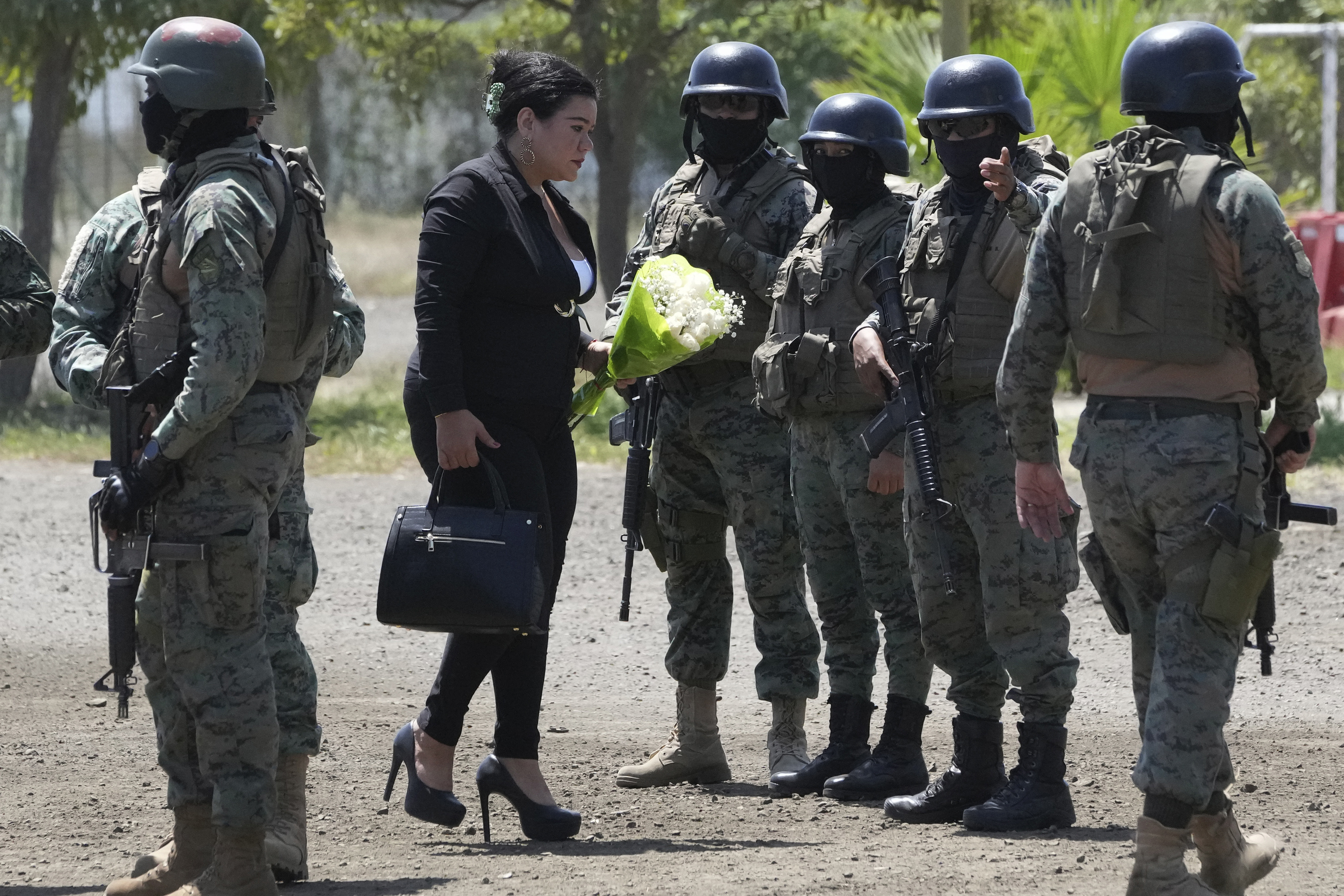 A woman, wearing black, brings a bouquet of white flowers. A line of armed guards in helmets stands in front of her.