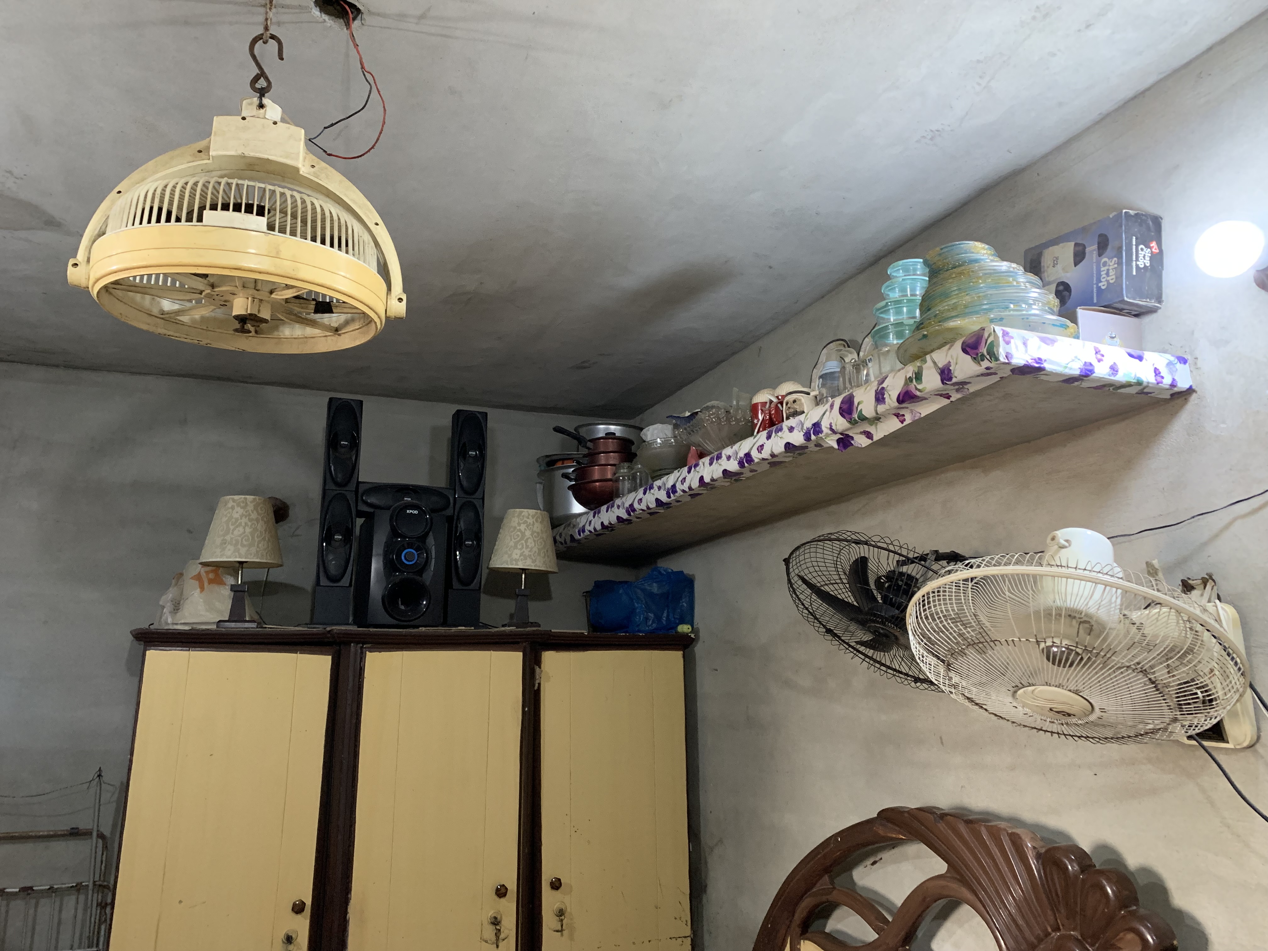 A photo of a shelf on a wall with two fans attached to the wall, below the shelf, with a wardrobe on the wall next to it with a variety of things on it including a lamp and a boombox.