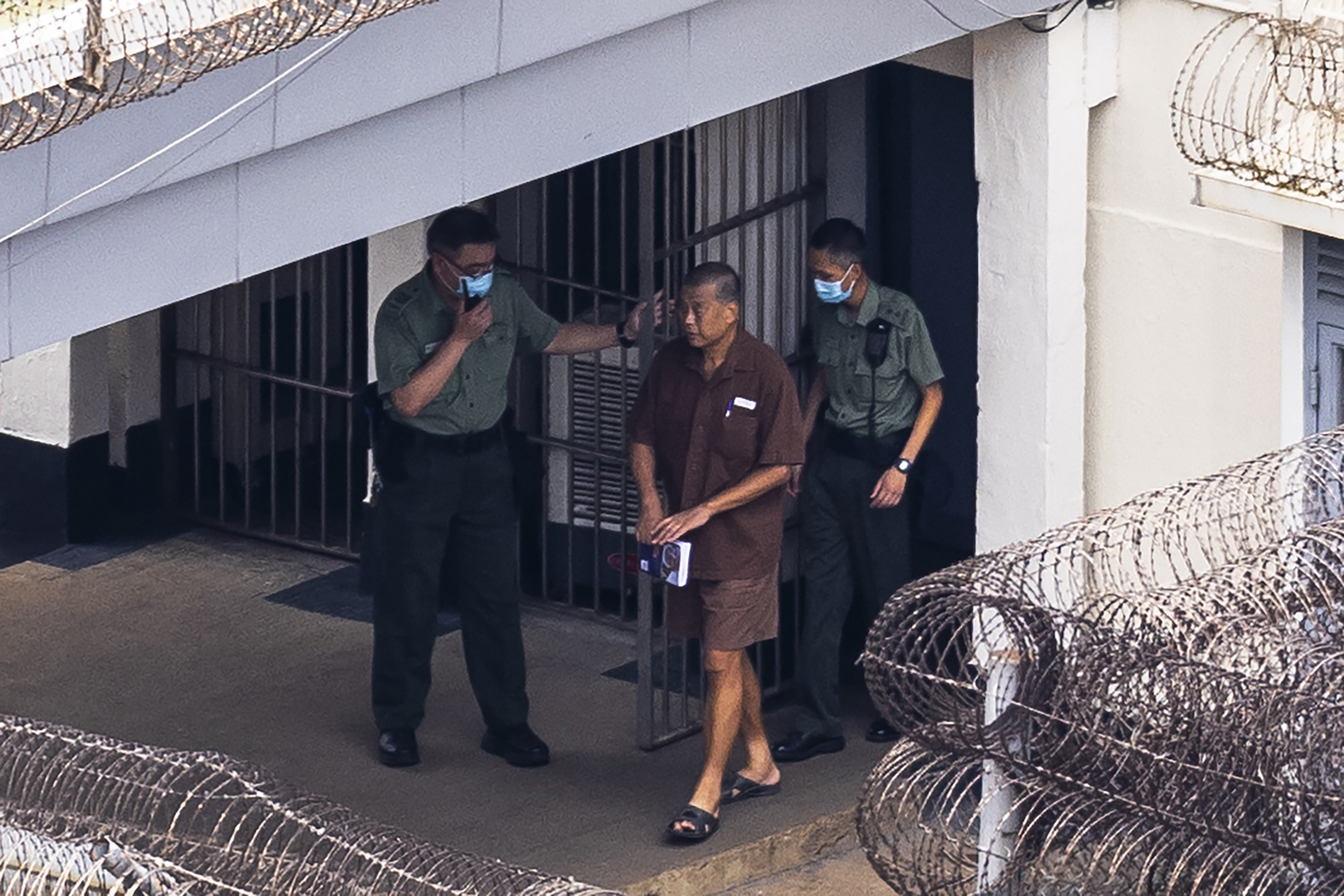 Jimmy Lai being escorted by two prison officers at Hong Kong's Stanley prison. The jail is a maximum security facility and there are coils of barbed wire on the top of the walls. Lai is wearing a brown short sleeved shirt, shorts and slides.