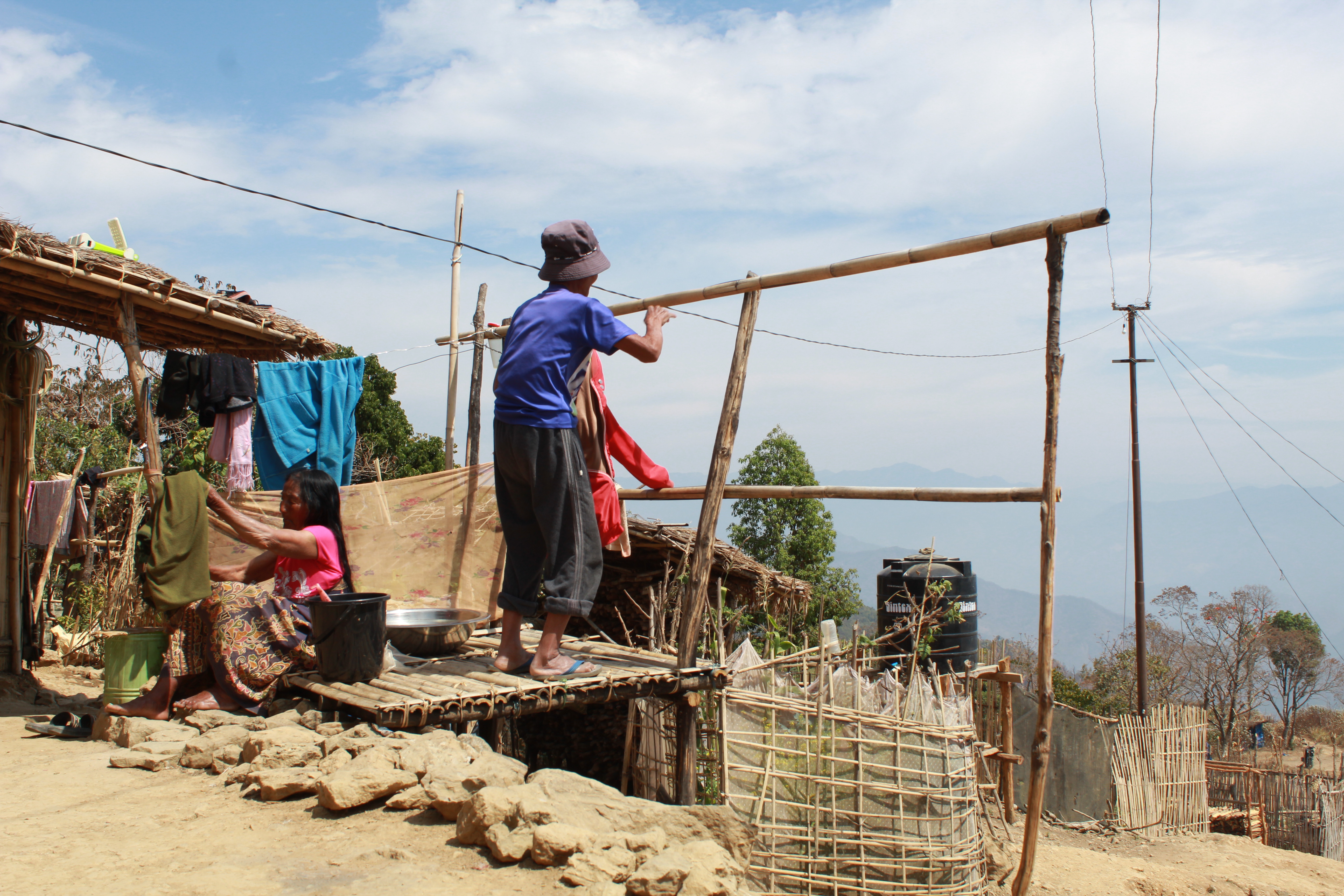 A couple hanging ot their laundry. He is standing and holding some clothing with a drying stand in front of him. She is sorting laundry from a bucket. They are wearing T-shorts and sarongs. He is wearing a bucket hat. It looks dry. Hills are visible in the background through haze