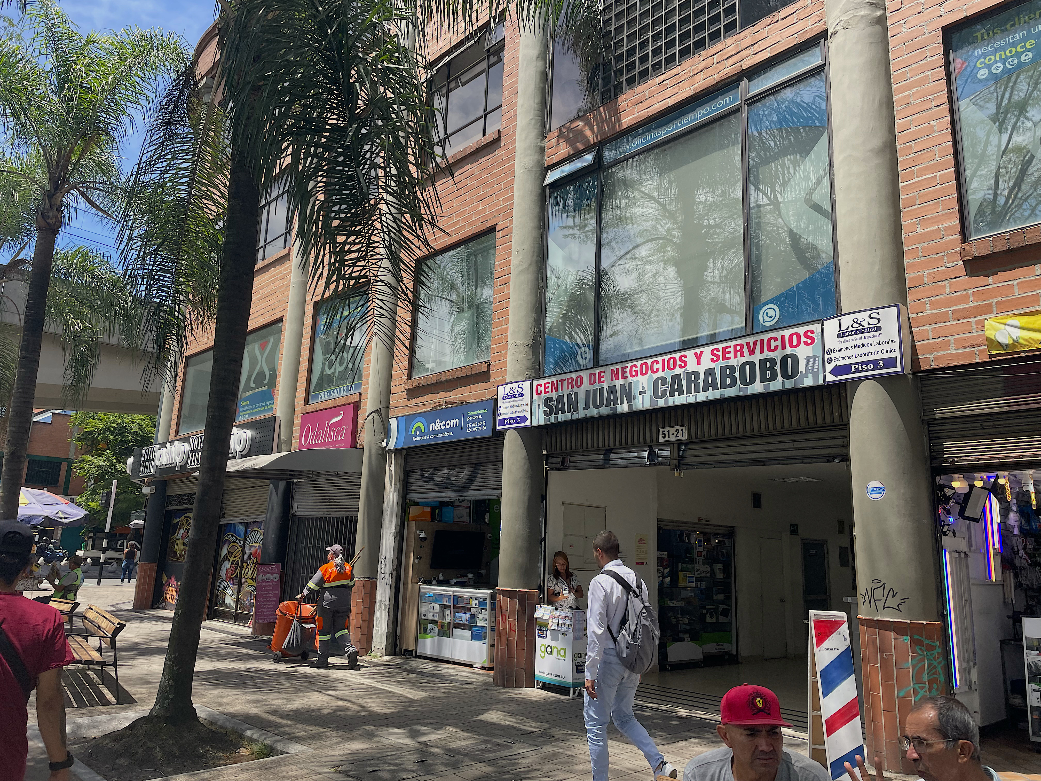 The exterior of a building in Medellin Colombia, that has a sign that reads: "Centro de Negocios y Servicios. San Juan-Carabobo." Palm trees are planted on the sidewalk outside.