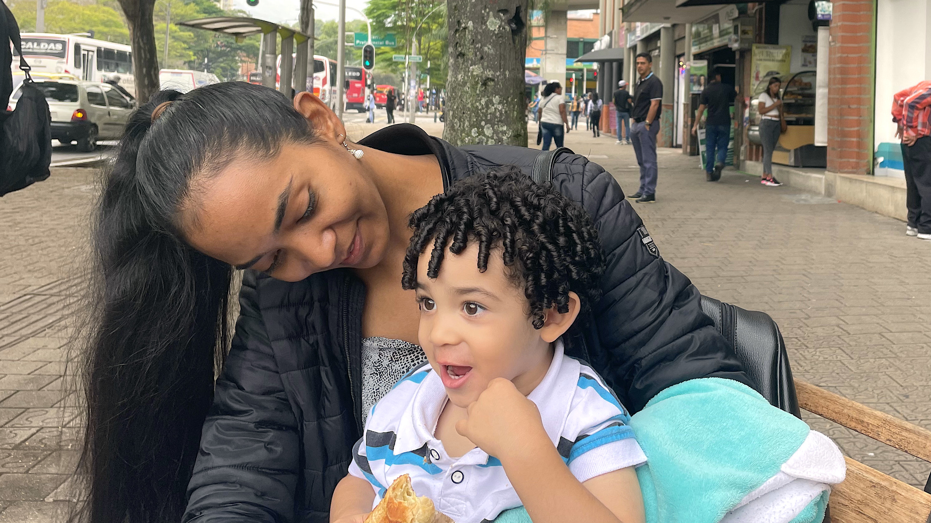 Yesica Cordoba sits outside on the sidewalk in Medellin, Colombia, with her young son perched on her knee. She leans over to look at him.