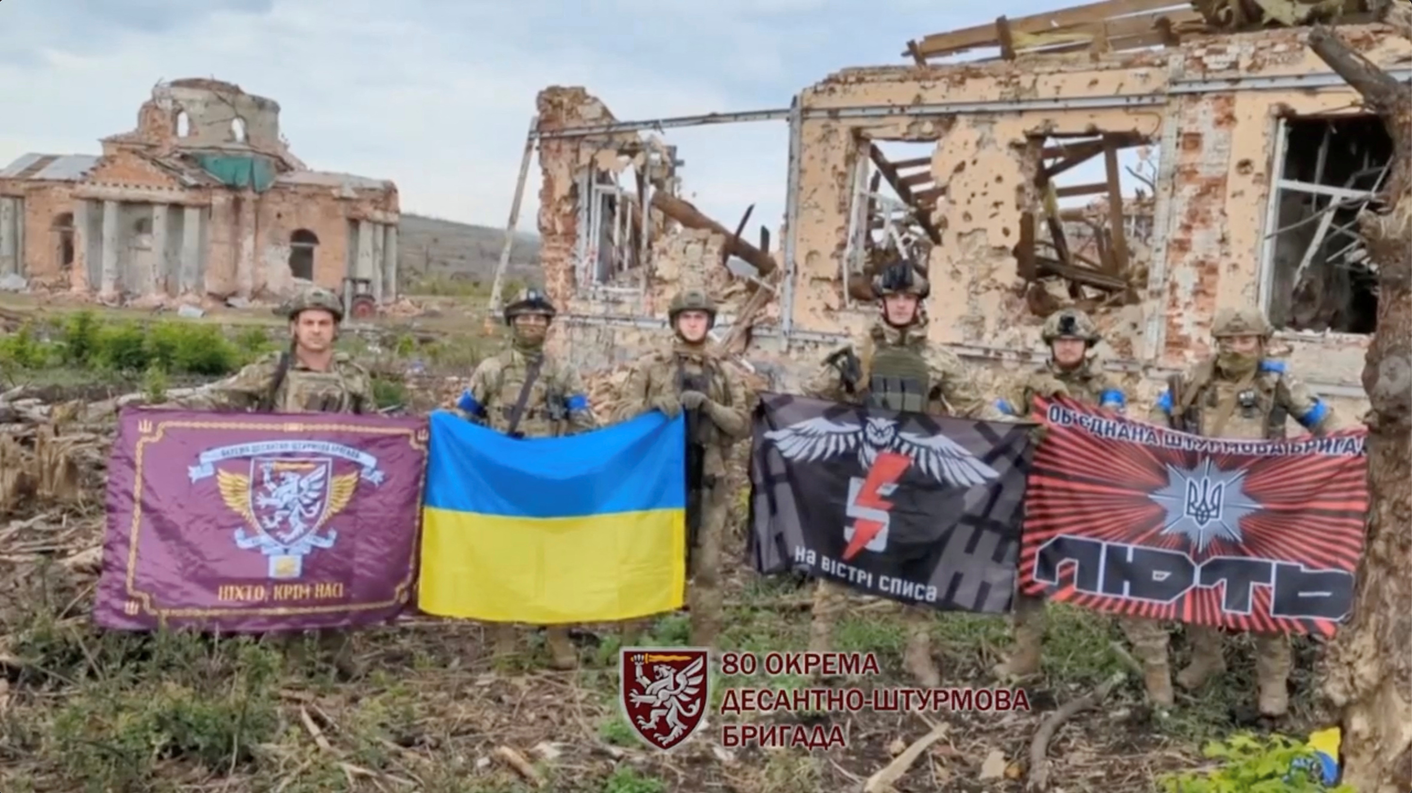 Soldiers hold the Ukraine flag and flags from their fighting groups in front of ruined buildings in Klishchiivka
