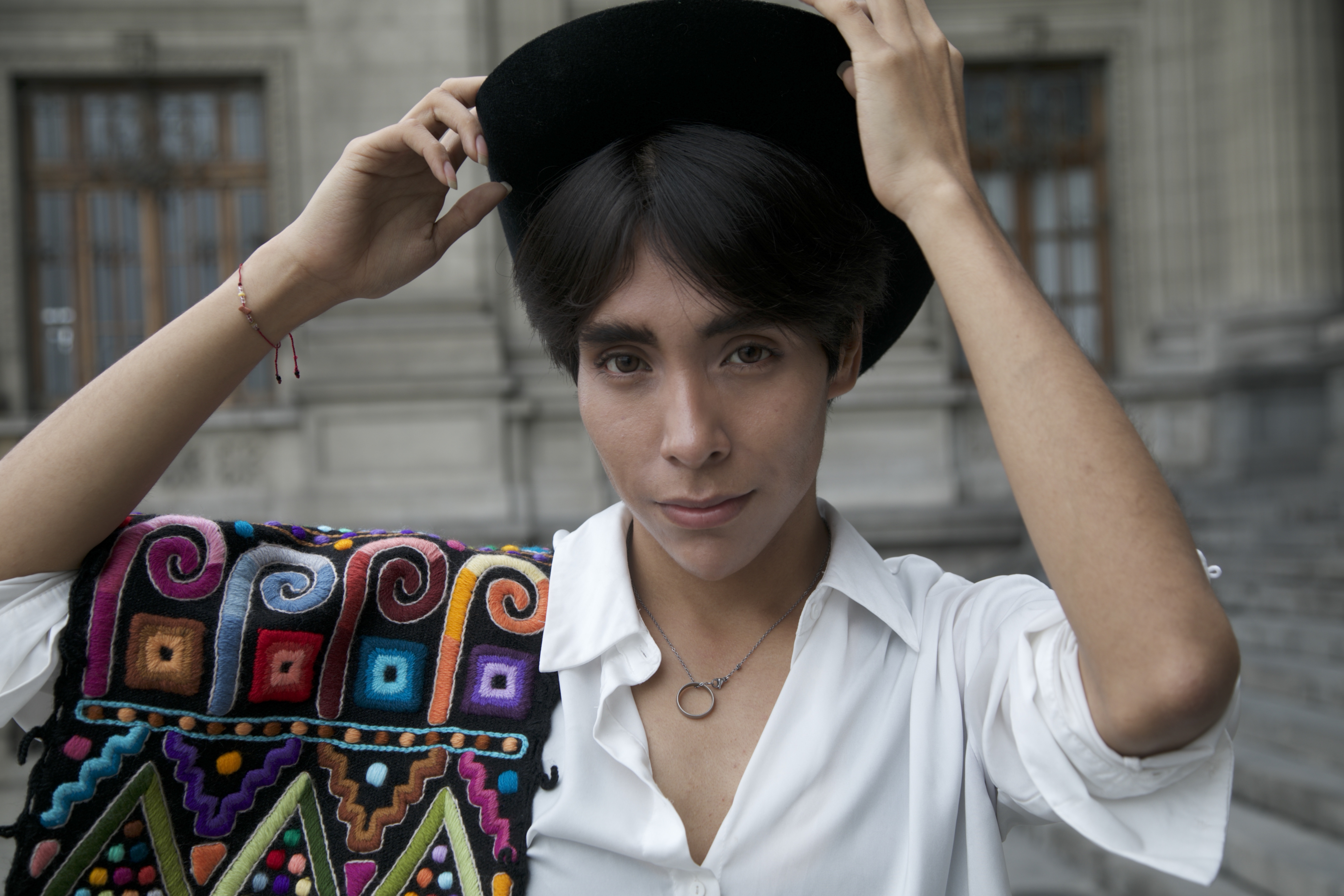 A young Peruvian man lifts his hands to hit hat as he poses for a photo. He wears a crisp white shirt, and over his shoulder is an Indigenous belt, colorfully embroidered.