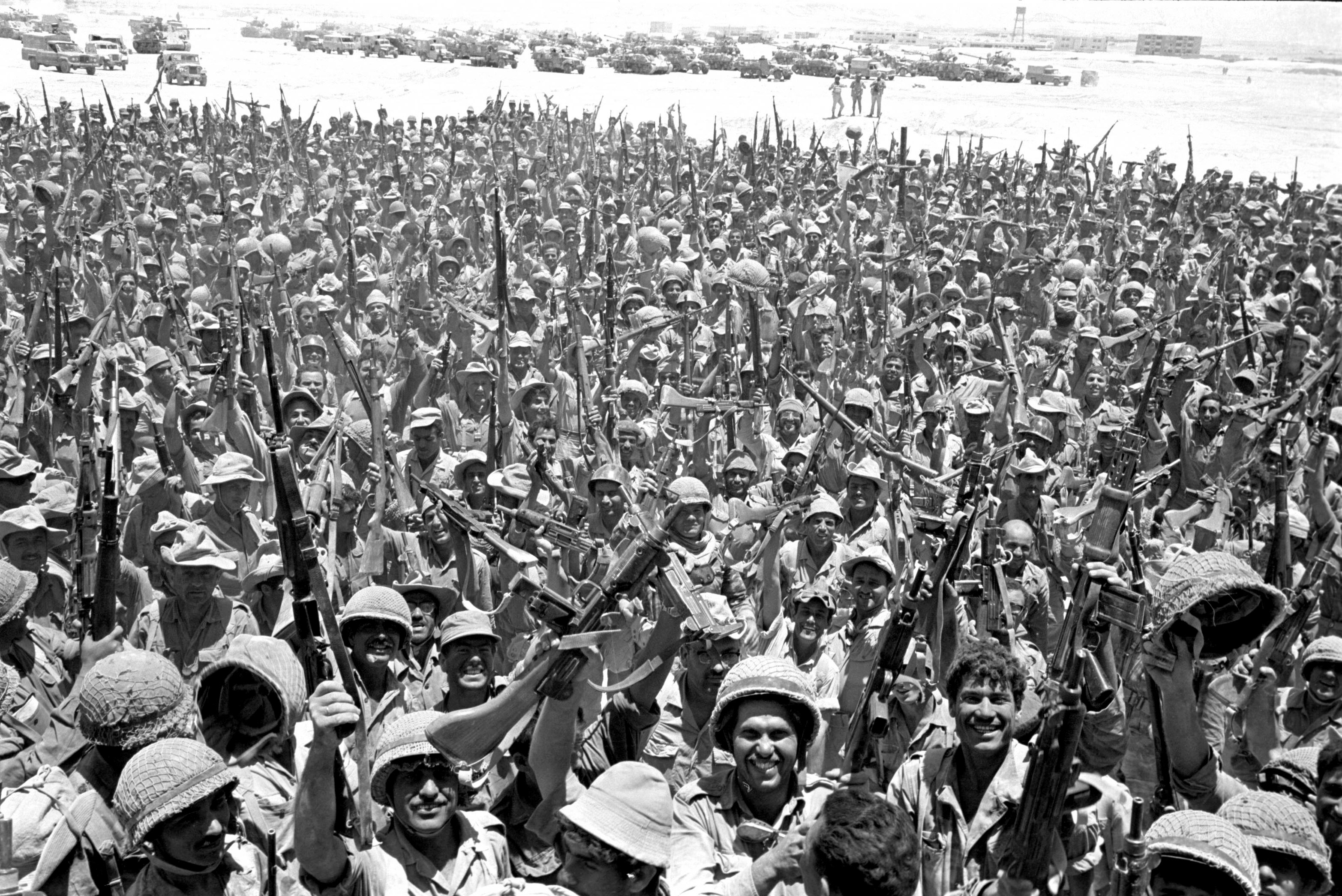 A black-and-white antique photo shows hundreds of Israeli soldiers in helmets and fatigues celebrating, lifting their rifles into the air.