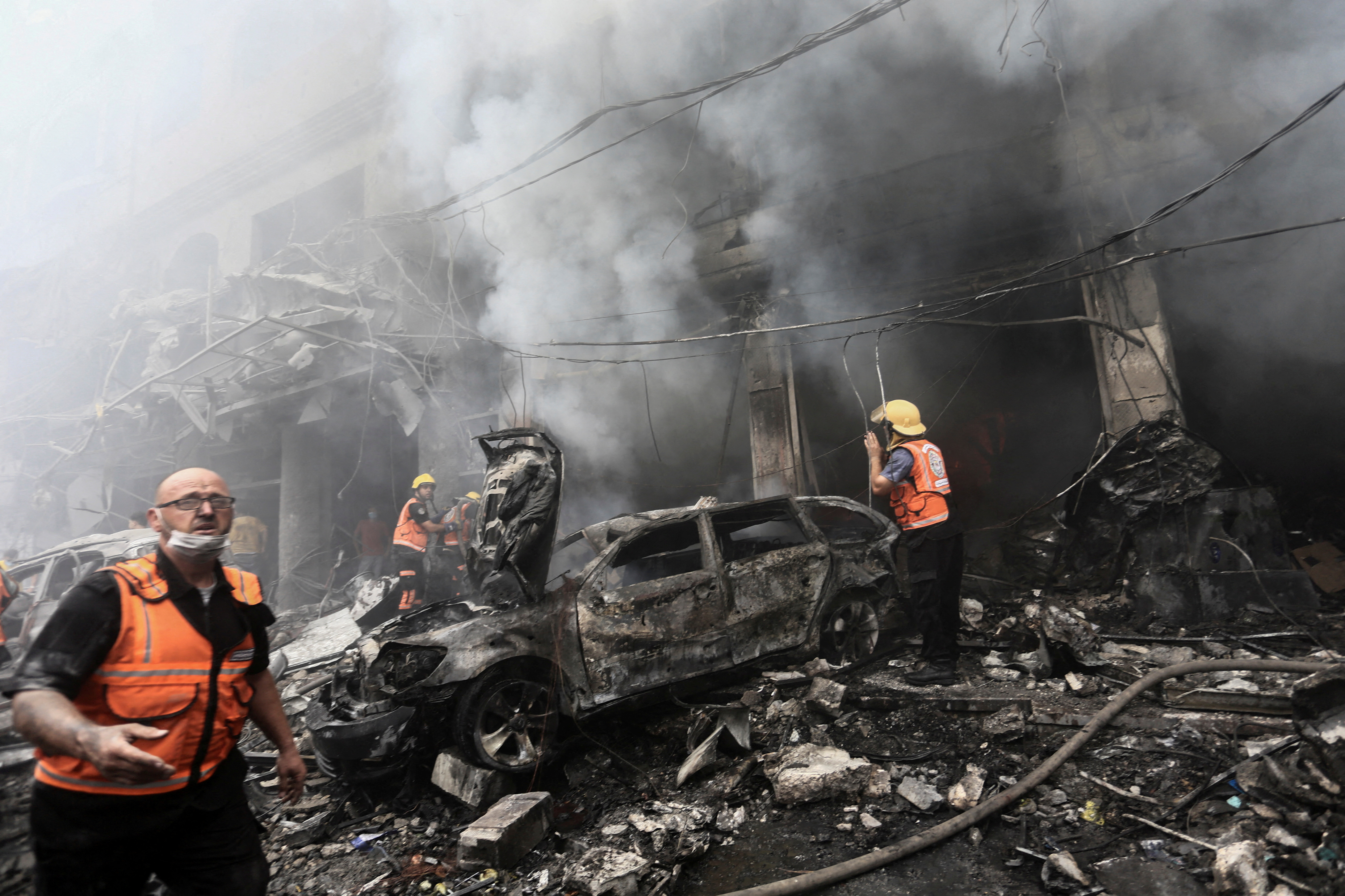 Clouds of smoke rise from a bombed-out refugee camp, as rescue workers approach a car charred by flames.