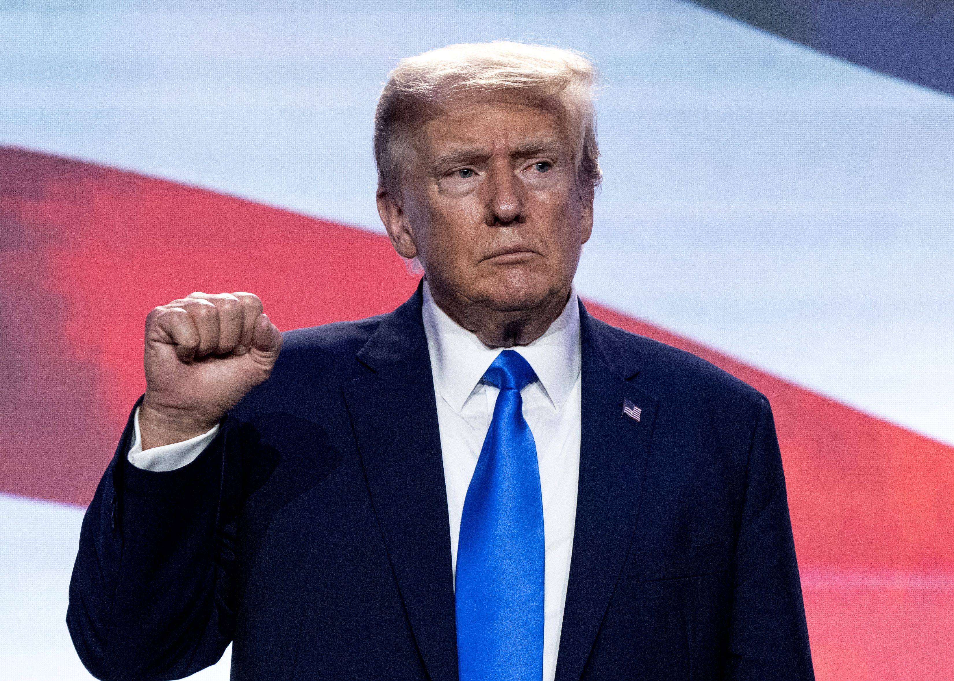 Trump standing in front of a red, white and blue backdrop, He is holding up his hand in a small fist.
