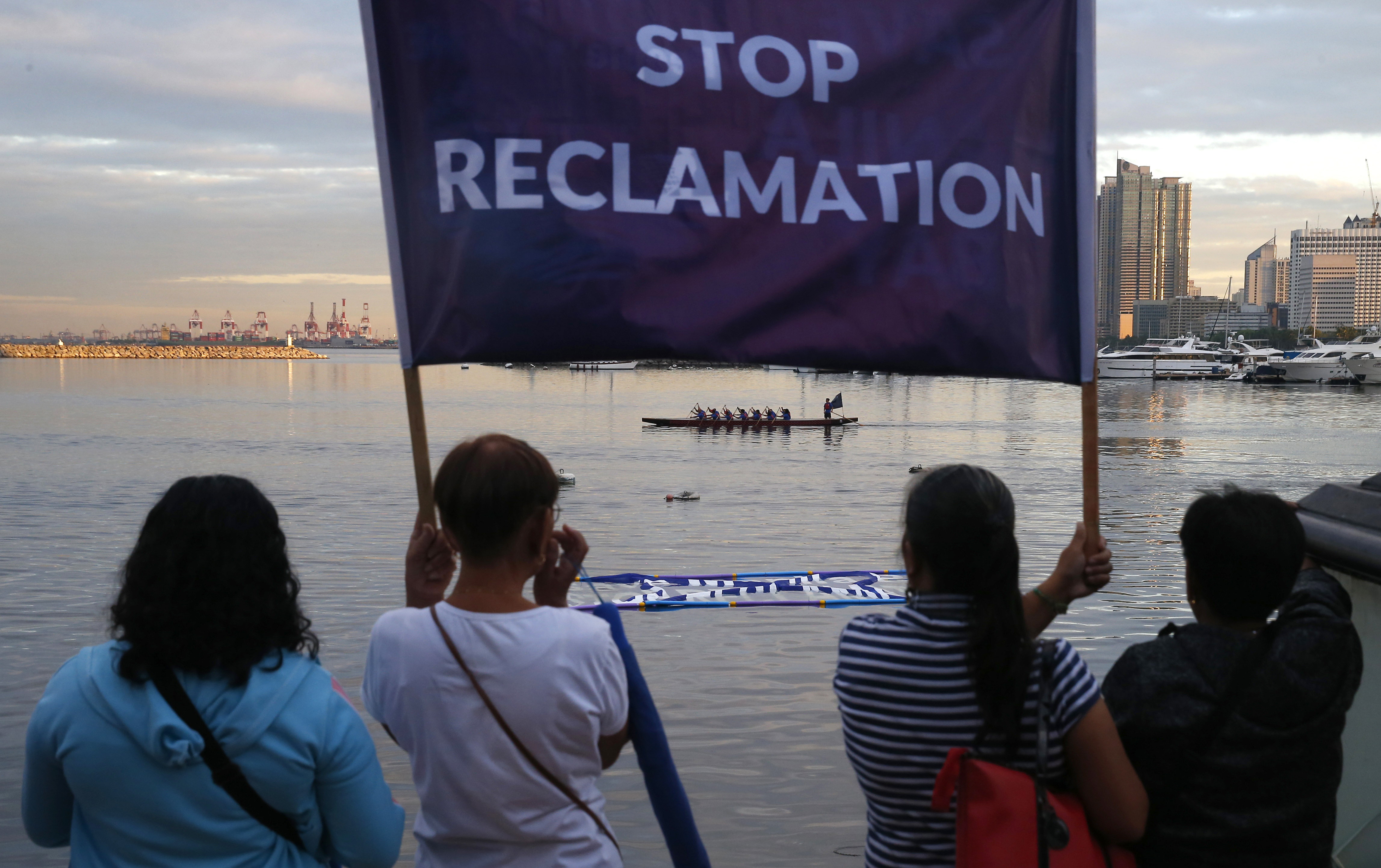 Protesters holding a banner reading 'Stop Reclamation'. They are looking out across the bay. There is a dragon boat crew on the water