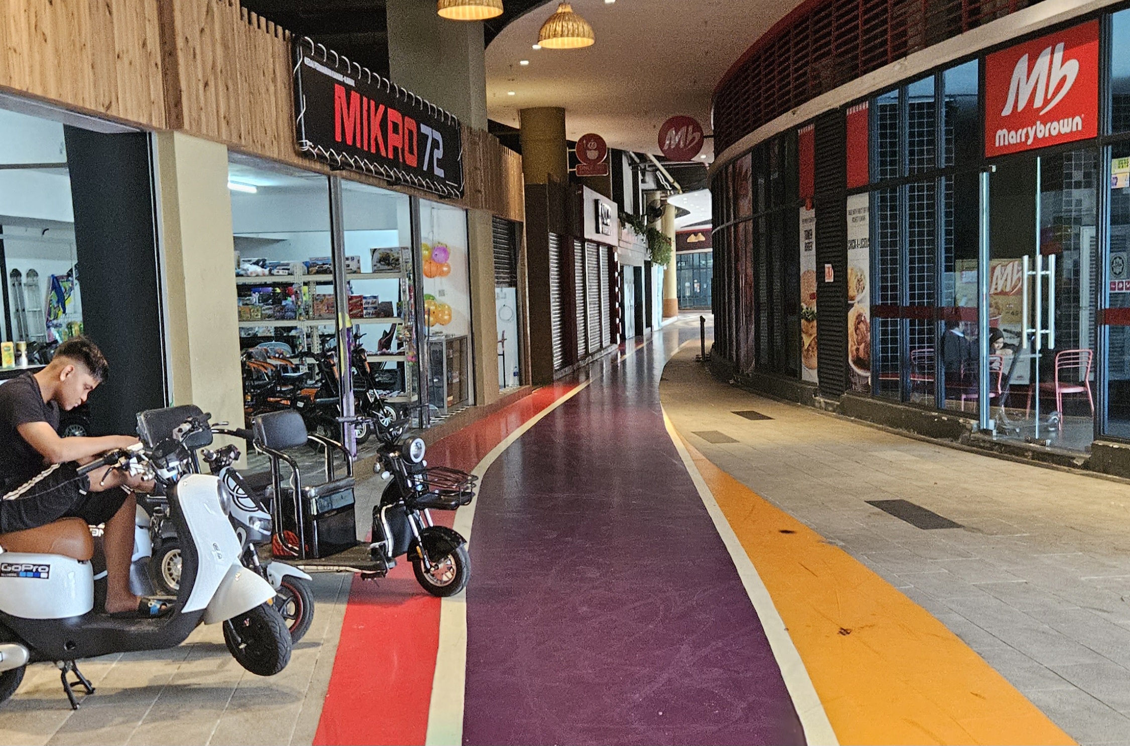 A man sitting on a scooter outside a shop that offers the bikes to rent. There is.a fast food outlet opposite but the area is deserted.